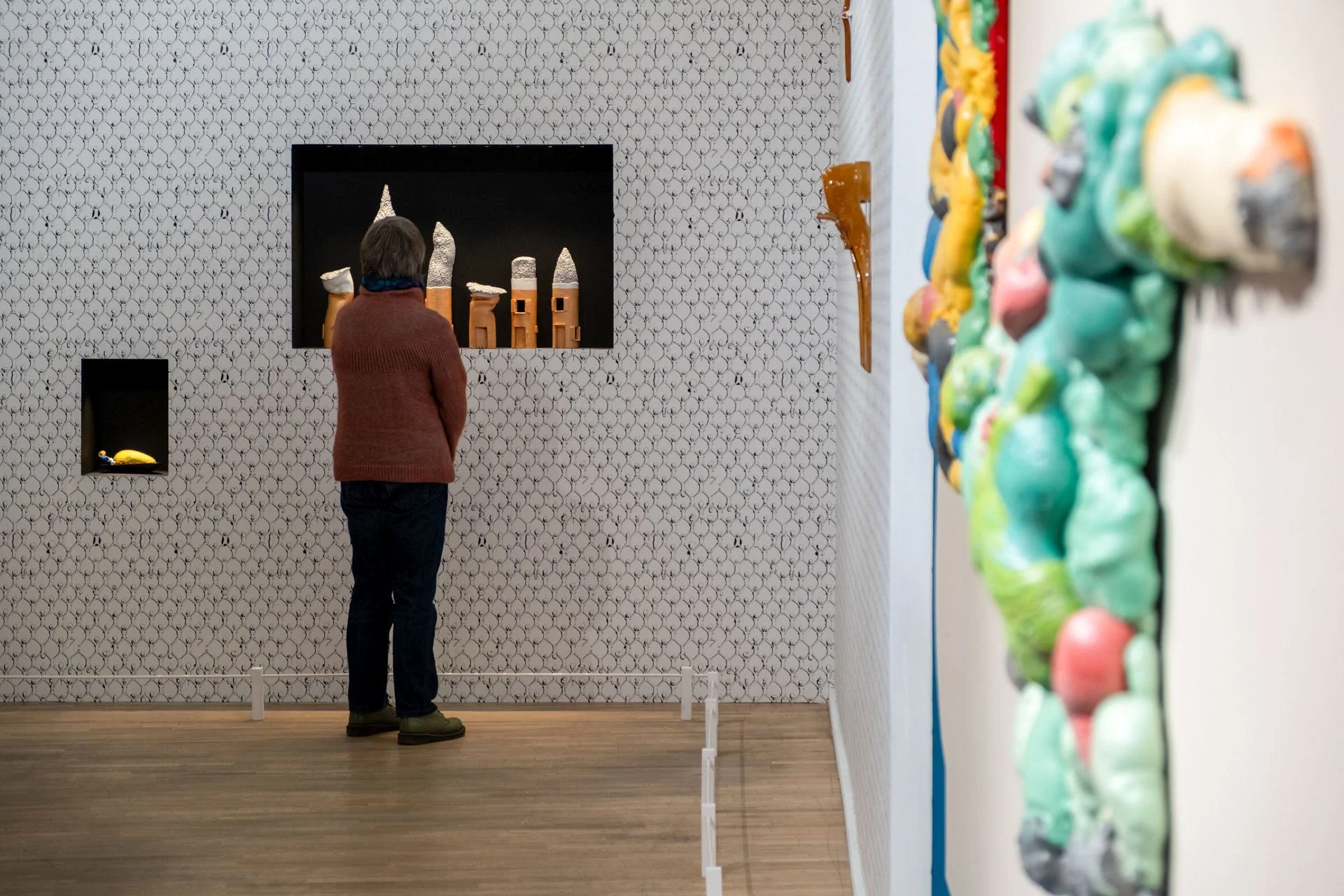 Une personne regarde des sculptures en céramique dans une galerie d'art, avec une mosaïque artistique sur le mur en arrière-plan et des œuvres colorées sur le côté droit.