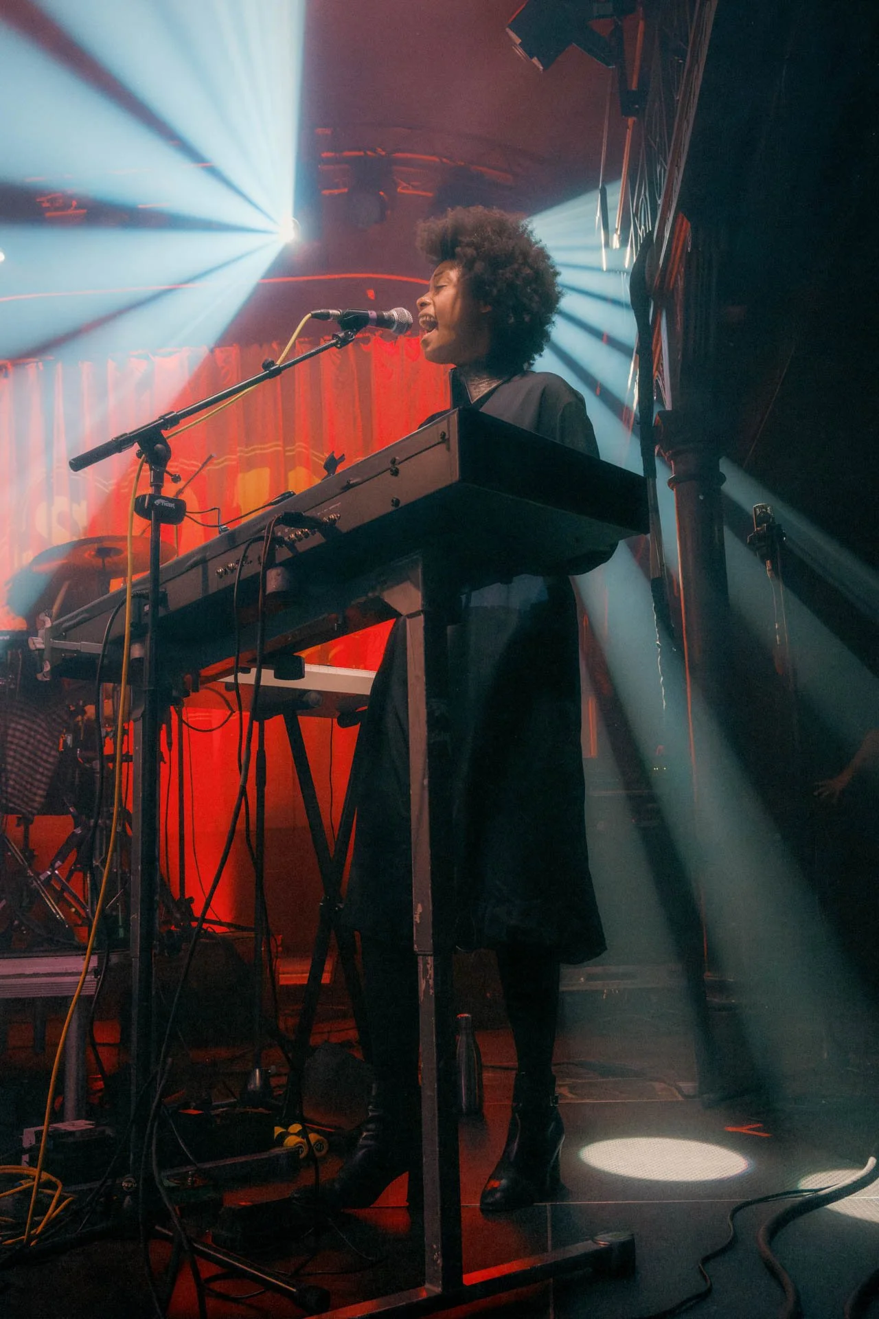 Une femme chante en jouant du clavier électrique sur scène, éclairée par des lumières de scène colorées et un faisceau lumineux blanc en arrière-plan.