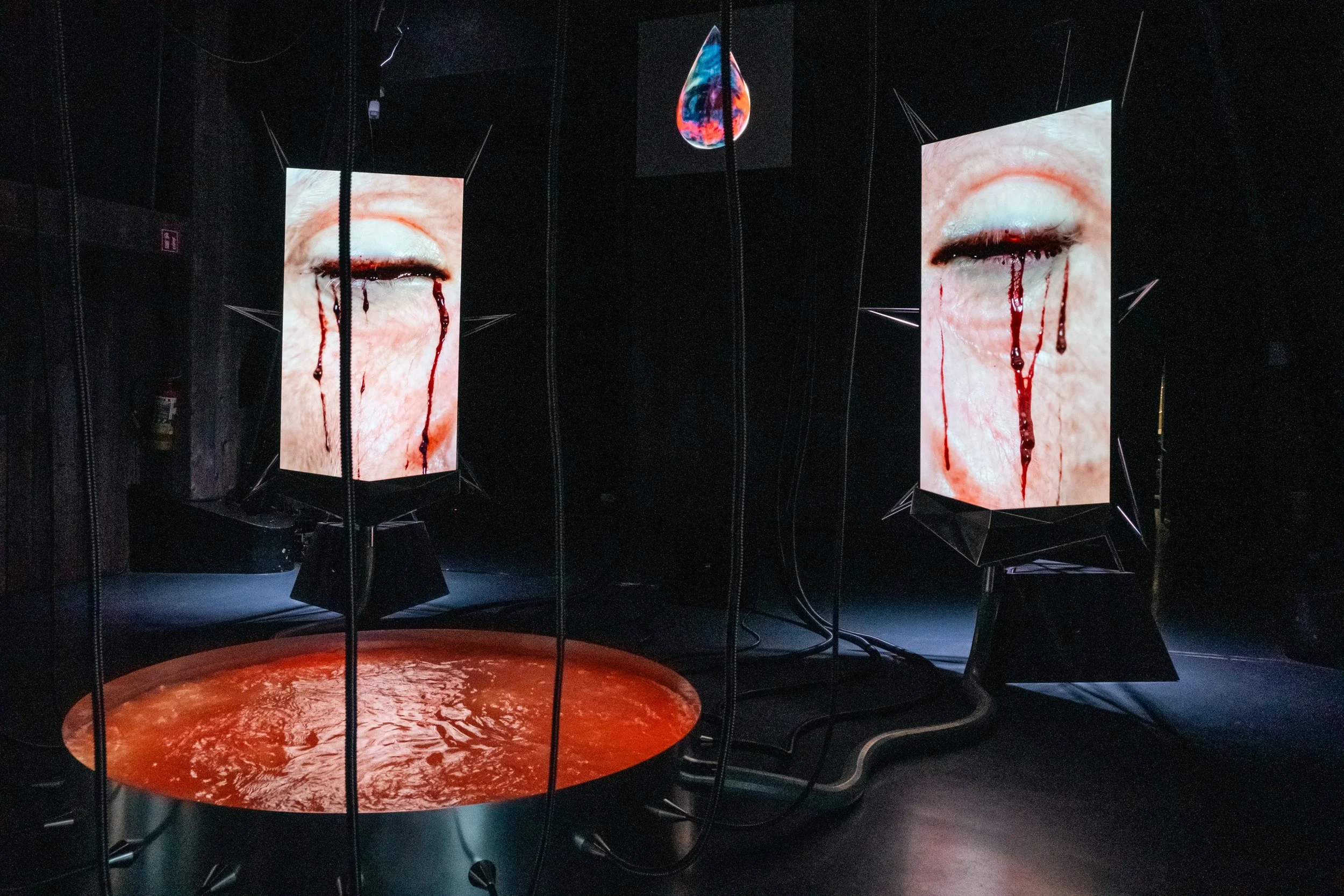 Installation artistique sombre avec plusieurs écrans affichant des images de yeux avec du sang coulant, une dalle verte euh en forme de goutte suspendue au centre avec des couleurs vives. L'ensemble crée une atmosphère intense et inquiétante.