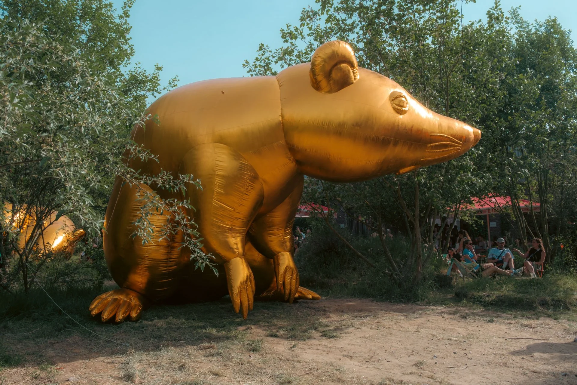 Statue géante d'une souris en ballon doré dans un parc, avec des arbres et des personnes assises en arrière-plan.