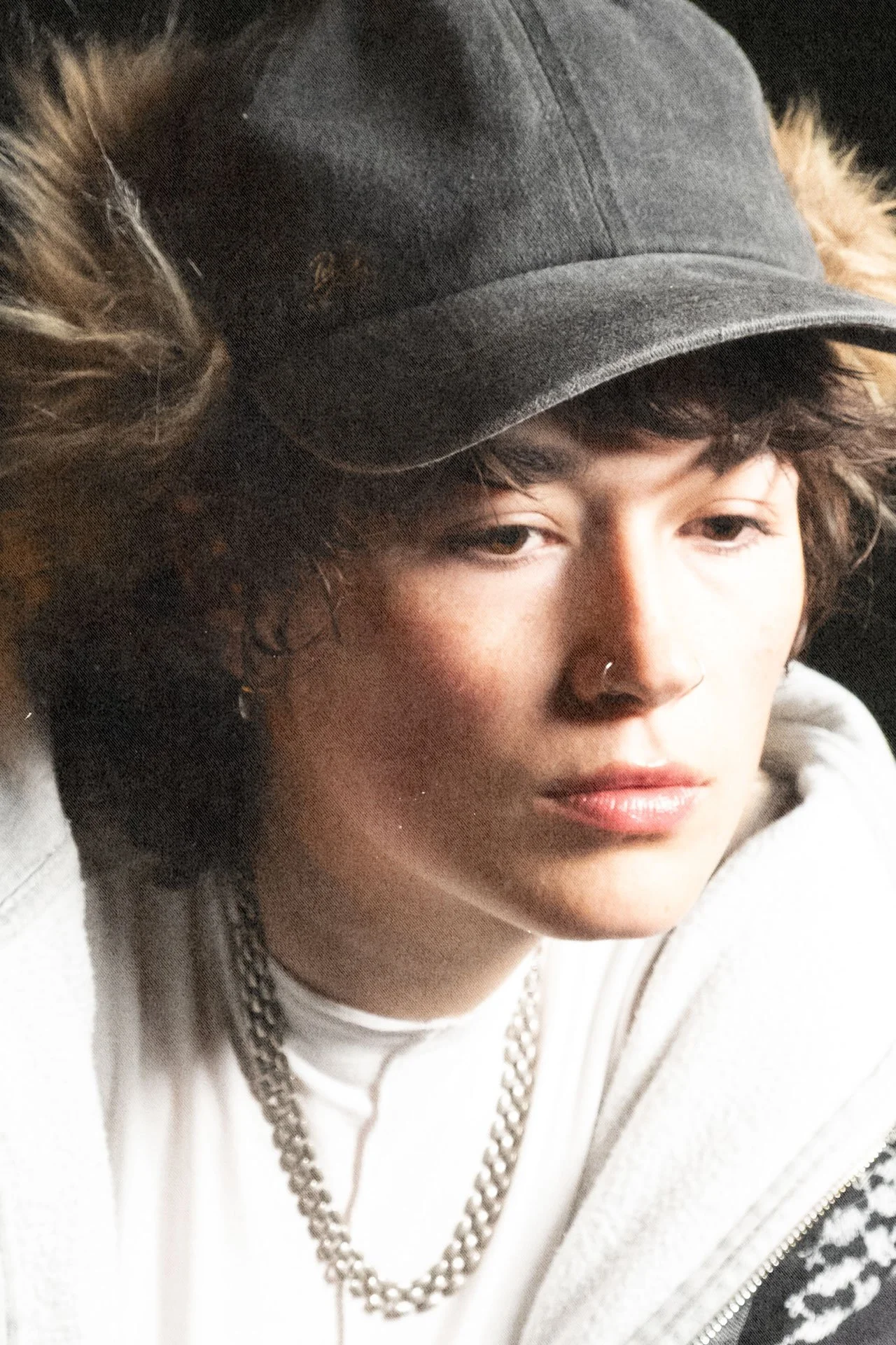 Portrait d'une jeune femme avec un chapeau gris, des cheveux bouclés, portant un collier en métal et une veste en fleece blanc avec des motifs noirs.
