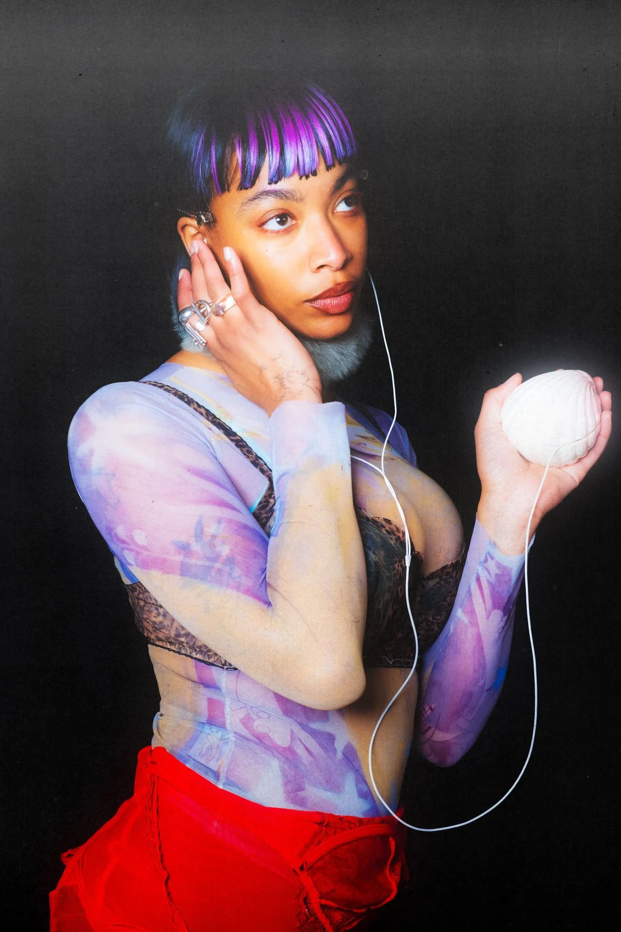 Une jeune femme avec des cheveux courts et teints en violet et noir, portant des vêtements moulants avec des motifs colorés, des bijoux en argent, et tenant une boule blanche dans sa main. Elle porte des écouteurs et se tient contre un fond noir.