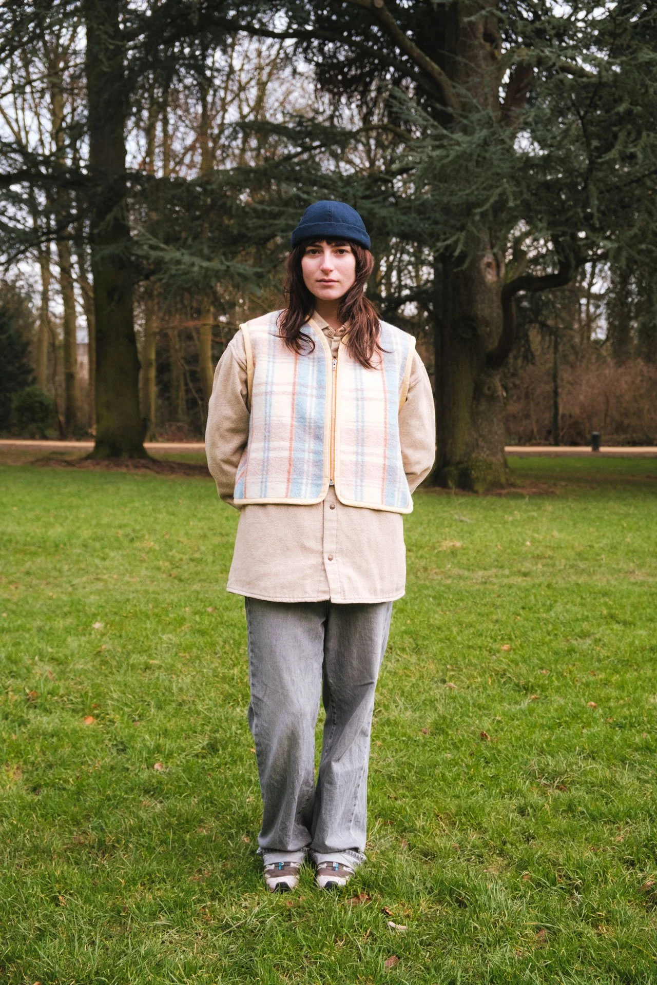 Jeune femme debout dans un parc verdoyant, portant un bonnet bleu, une veste beige à carreaux, un t-shirt beige, un pantalon gris et des chaussures de sport.