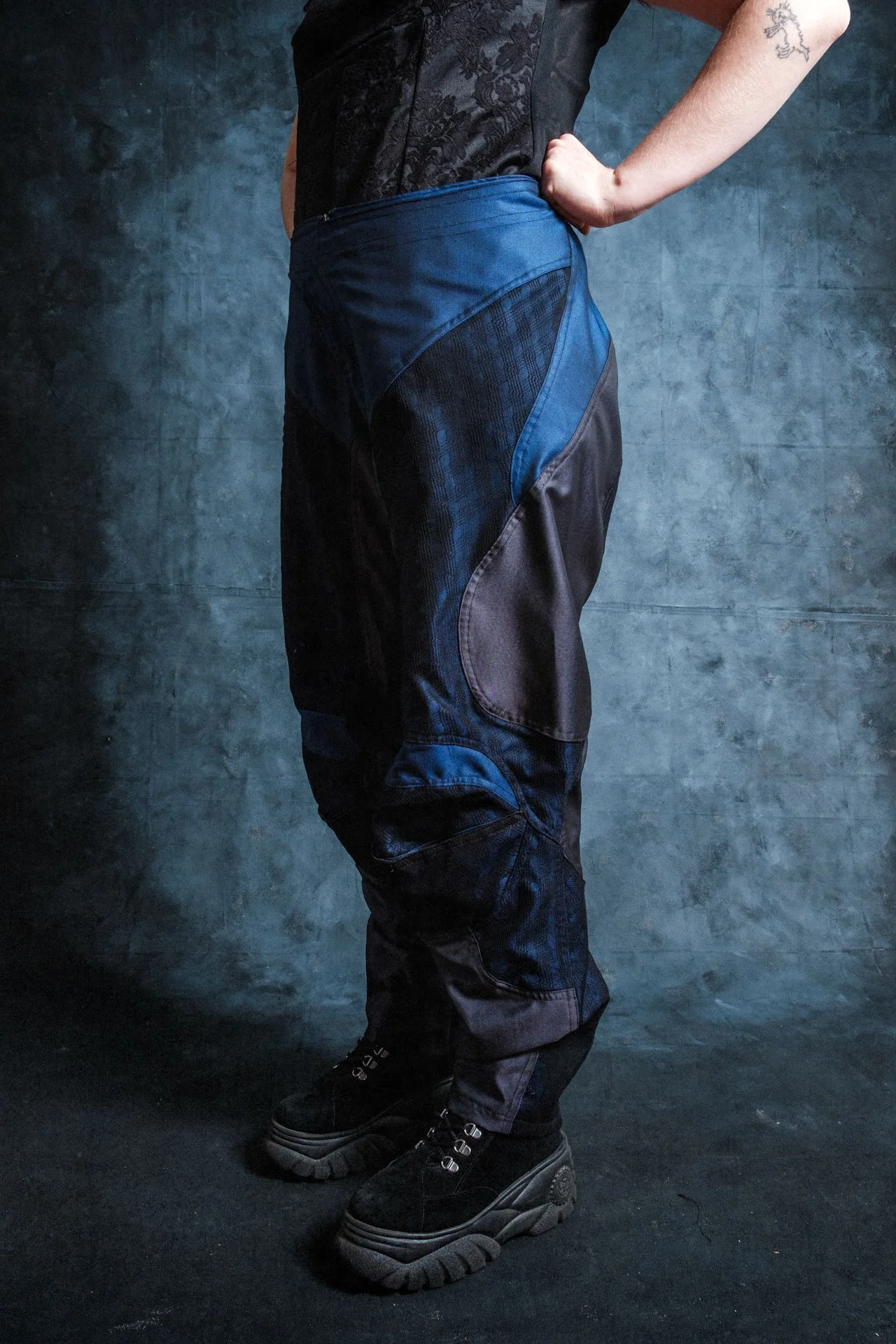 Membre d'une personne portant un pantalon de style utilitaire ou de sport, avec des chaussures noires épaisses, contre un mur gris foncé. La personne a le bras plié derrière le dos, et une tatouage est visible sur le bras droit.