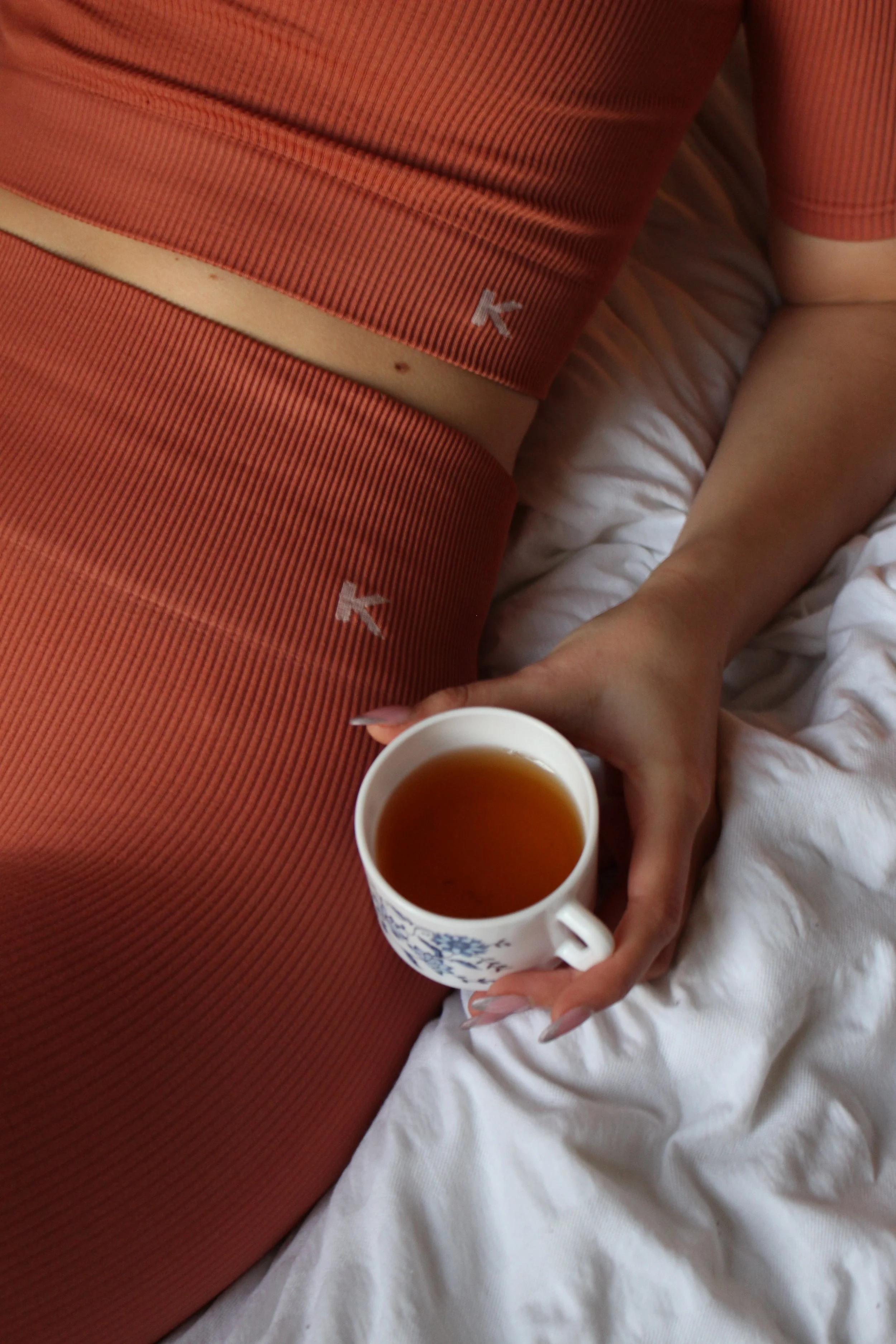 Une personne tient une tasse de thé ou d'une boisson chaude dans la main, assise sur un lit avec des draps blancs. La personne porte un haut orange avec une lettre 'K' en blanc. La photo est prise en gros plan, avec un focus sur la tasse et la main.