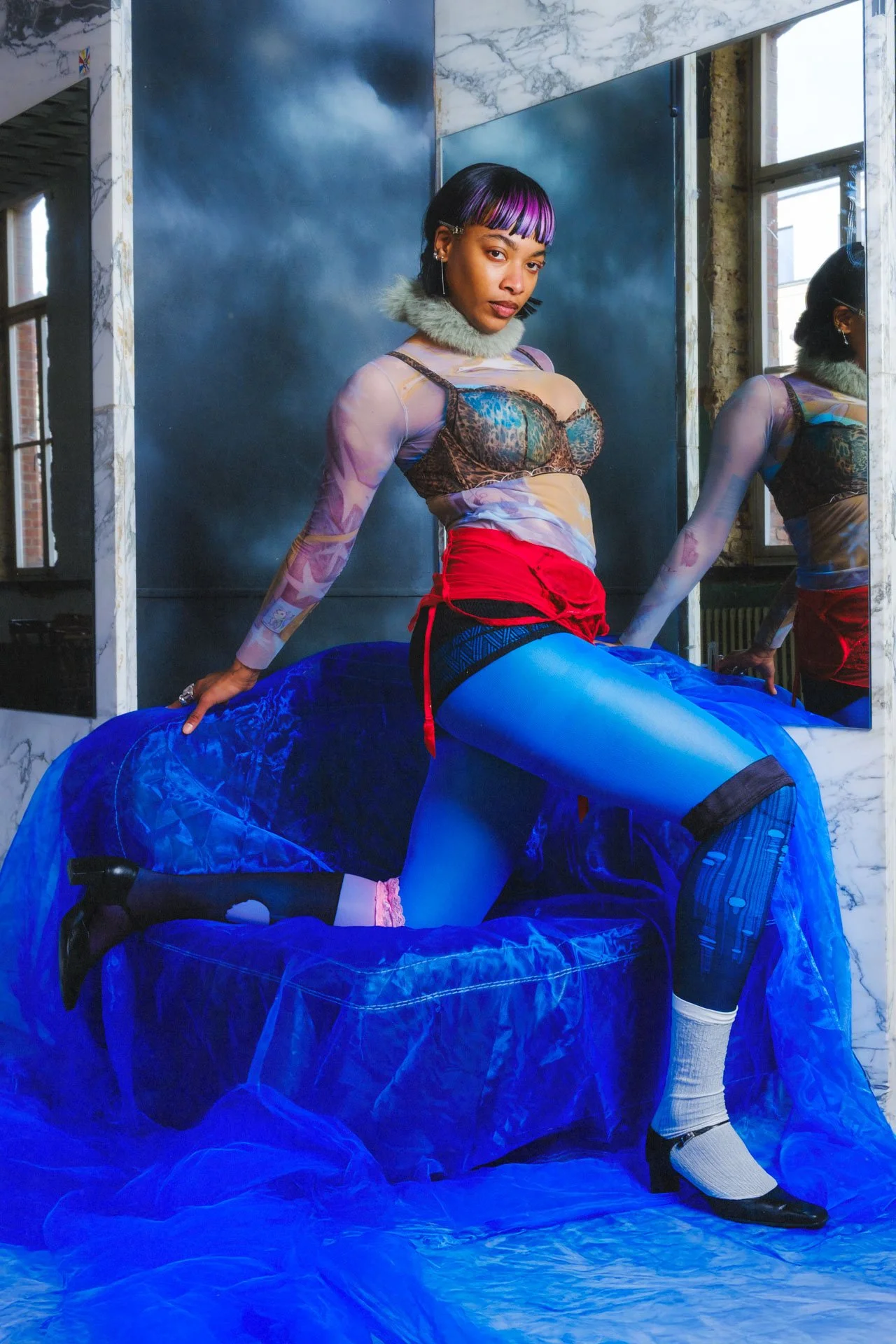 Une femme pose dans une pièce avec un décor urbain, portant un haut moulant en tissus transparent à motifs, une jupe courte rouge, des collants bleus, et des chaussures à talons noirs et blancs. Elle se tient sur un canapé bleu, avec un grand miroir 