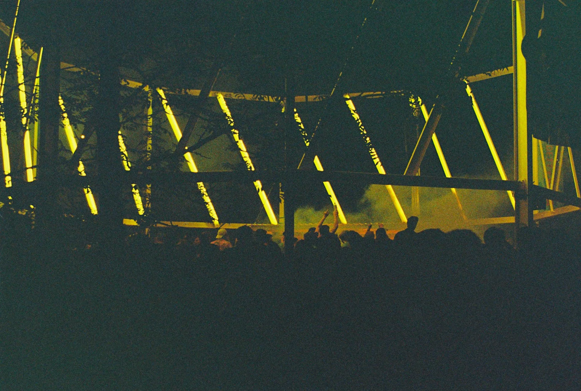 Une grande structure métallique illuminée en jaune, avec des personnes assises ou debout en dessous, lors d'une manifestation ou d'un spectacle nocturne.