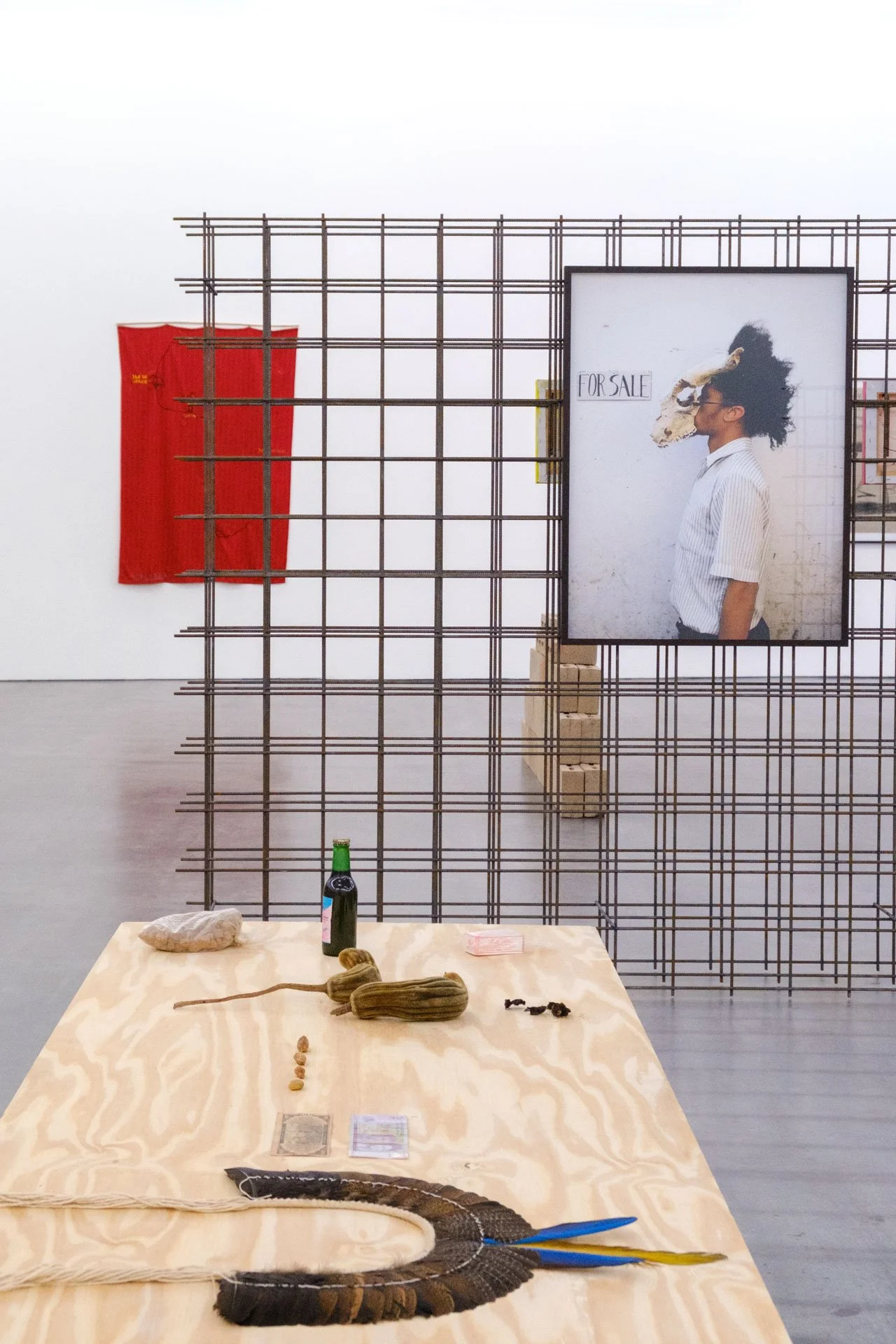 Une installation d'art contemporain avec une grille en métal, une affiche représentant une personne avec un masque animal, un drapeau rouge, une bouteille et des objets variés sur une table en bois.