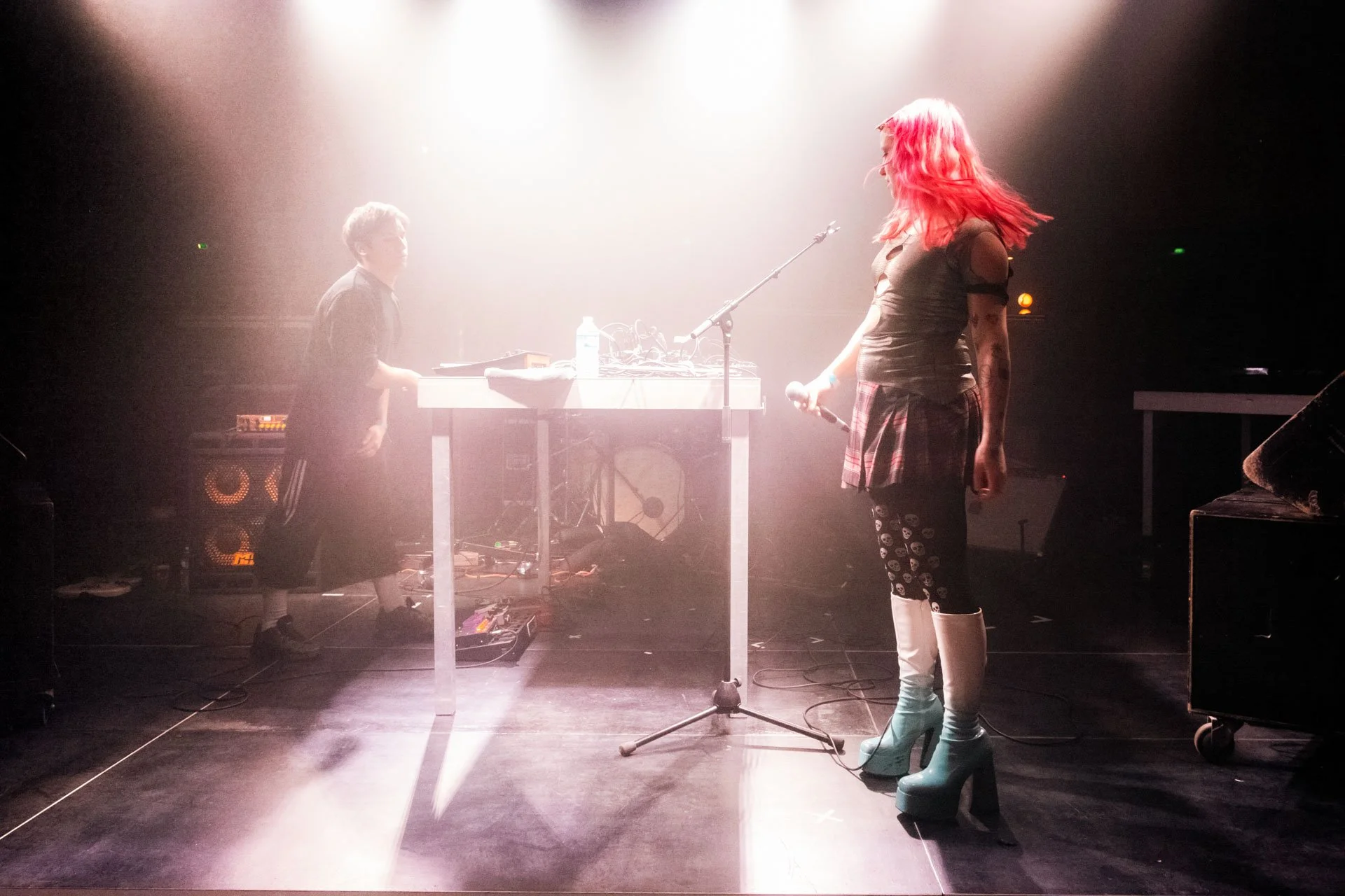 Une scène de concert avec une femme aux cheveux rouges, portant des bottes turquoise, et un homme à gauche derrière une table de DJ ou de musique, sous un éclairage tamisé.