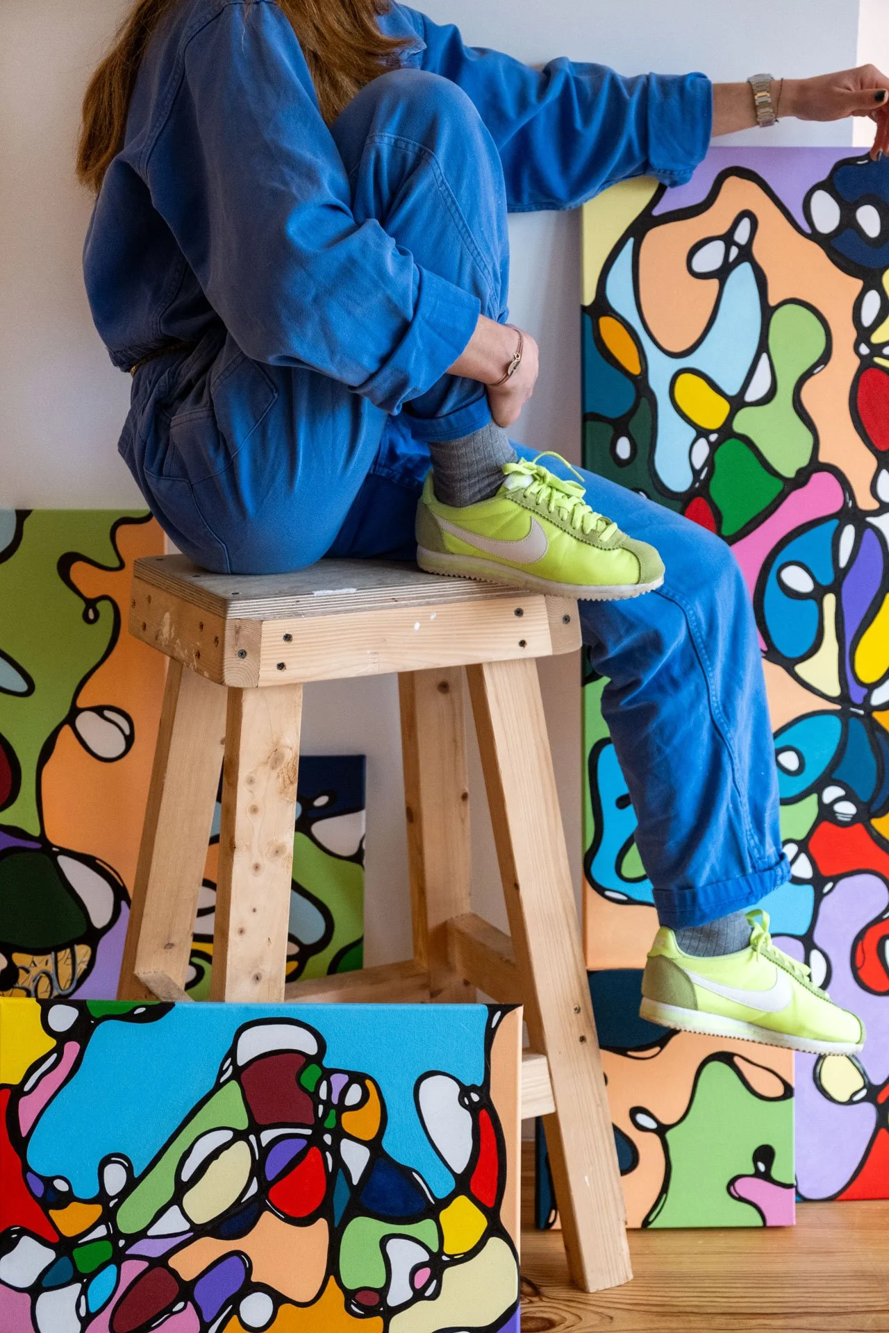 Une personne assise sur un tabouret en bois, portant une veste et un jean bleu, avec des baskets jaunes lime, devant un mur avec des œuvres d'art abstraites colorées.