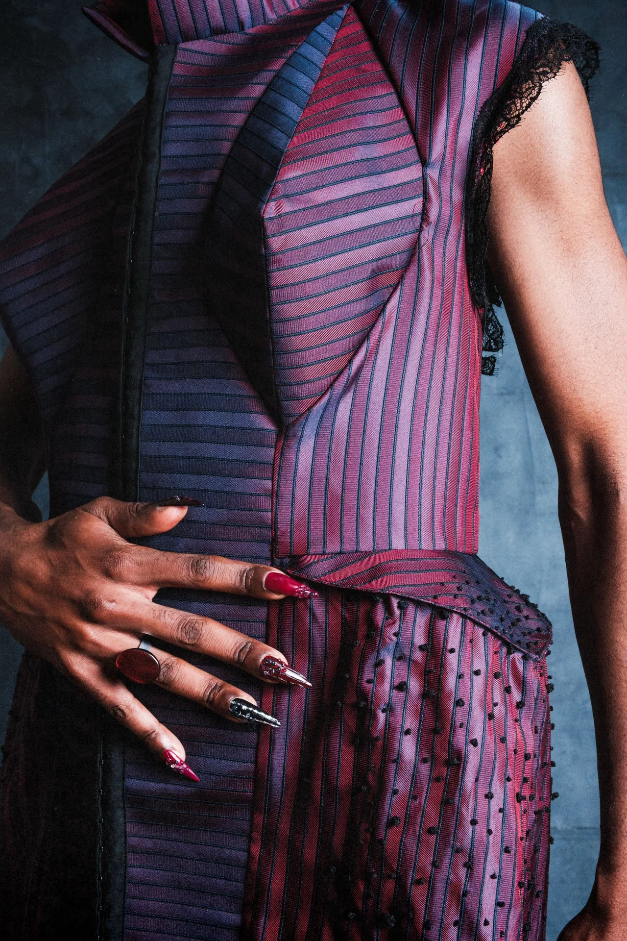 Une femme porte une robe sexy en tissu rayé violet et noir, avec des détails en dentelle noire. Sa main gauche repose sur sa hanche, montrant des ongles décorés de manière élaborée avec des motifs métalliques et rouges. Elle porte également une bague