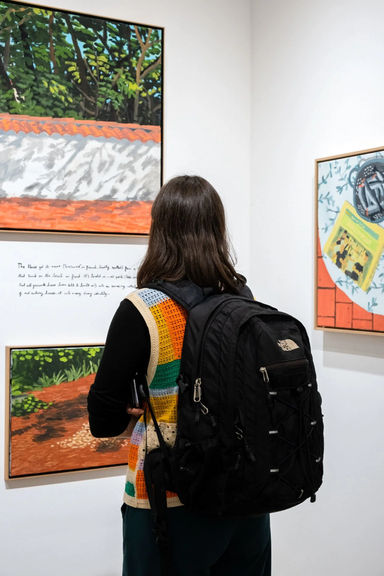 Une femme avec un sac à dos noir regarde une œuvre d'art dans une galerie, des tableaux colorés sont affichés sur le mur blanc.