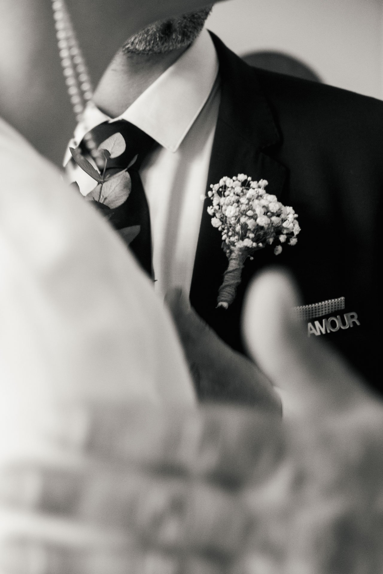 Une personne habillée en costume avec une épinglette de fleurs portée sur la poitrine, portant une boutonnière de fleurs, portant éventuellement une veste noire, avec un badge ou une étiquette partiellement visible avec le mot 'AMOUR'.