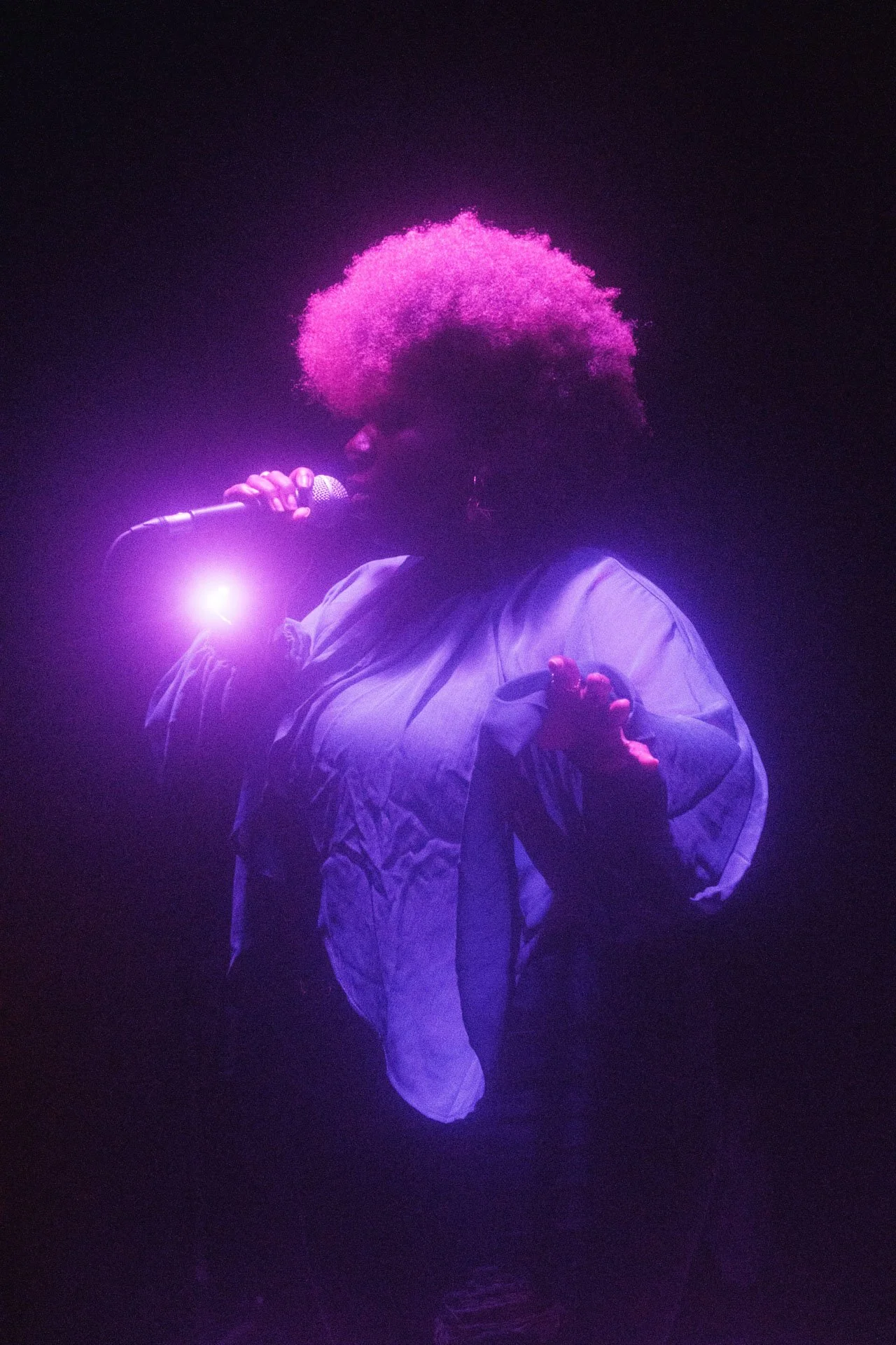 Une personne chante dans une lumière violette, portant une veste ample et tenant un microphone, avec un fond sombre.