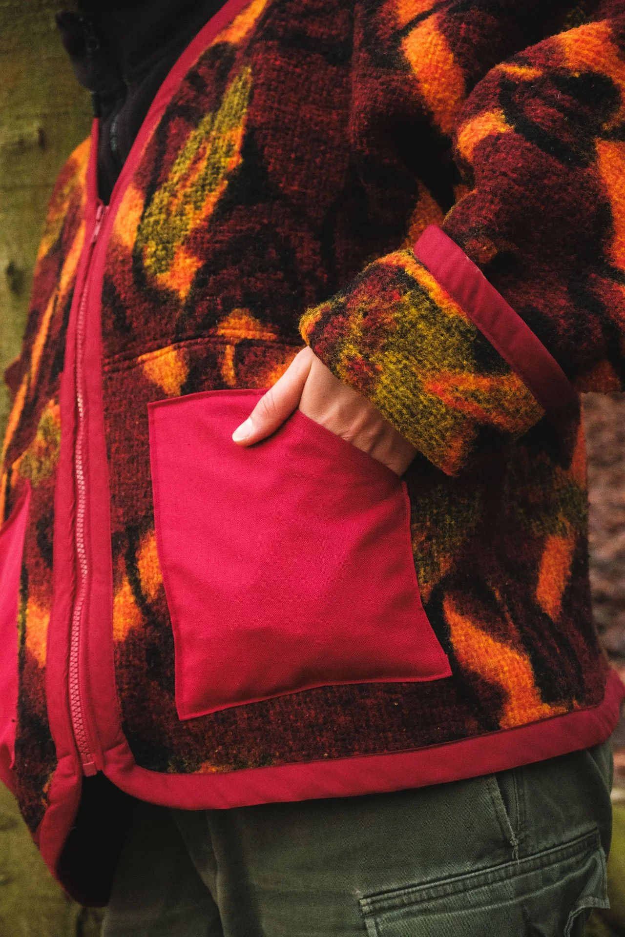 Une personne porte un manteau en laine à motifs orange, rouge, vert et noir avec un grand col, et une poche rouge à la poitrine. La personne a la main dans la poche.