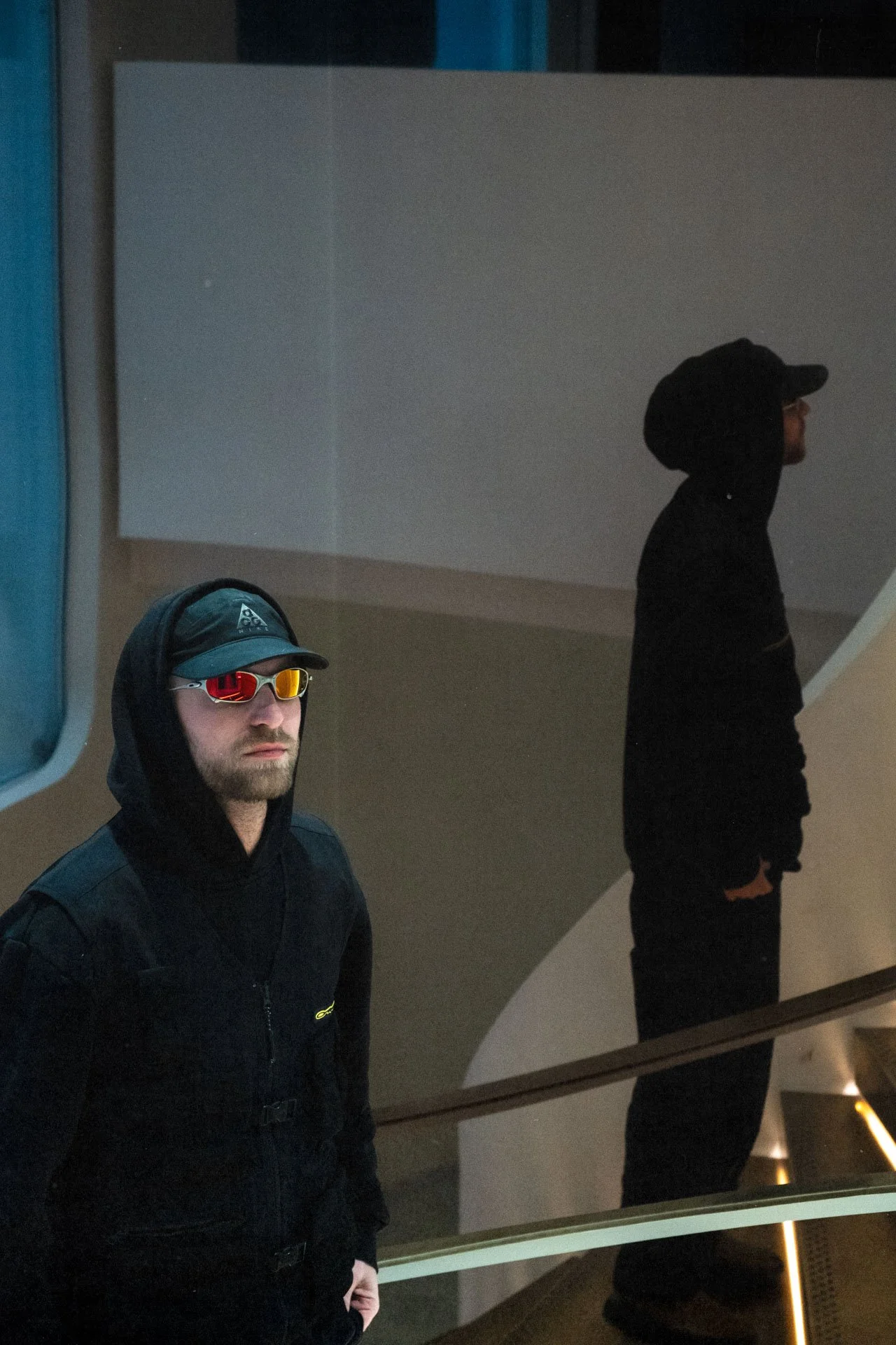 Un homme portant un chapeau, des lunettes de soleil rouges et une veste noire, regardant vers l'objectif, avec ses reflets visibles dans une surface vitrée derrière lui.
