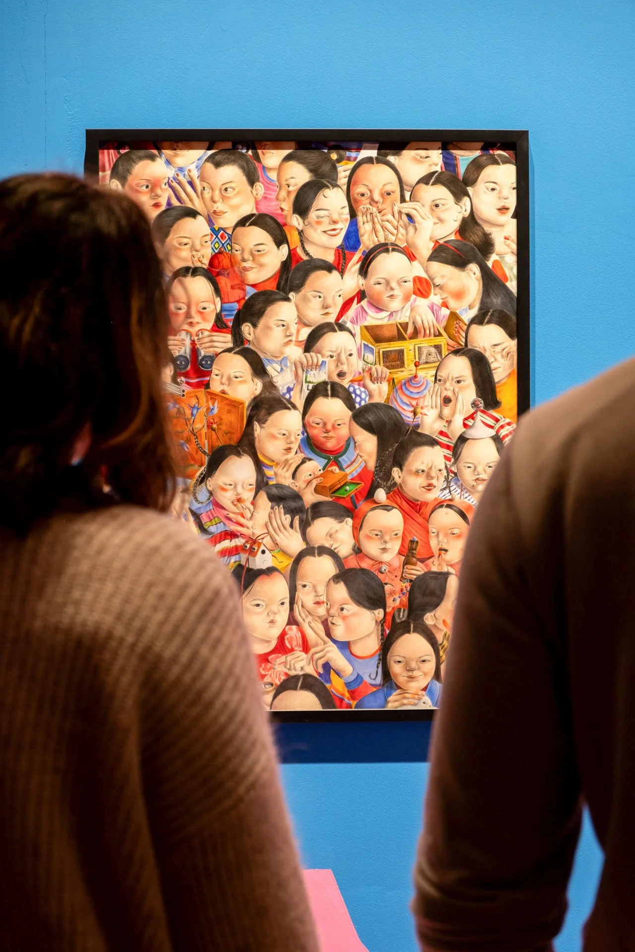 Deux personnes regardent une œuvre de art colorée représentant de nombreuses visages d'enfants avec diverses expressions, encadrée et exposée sur un mur bleu.
