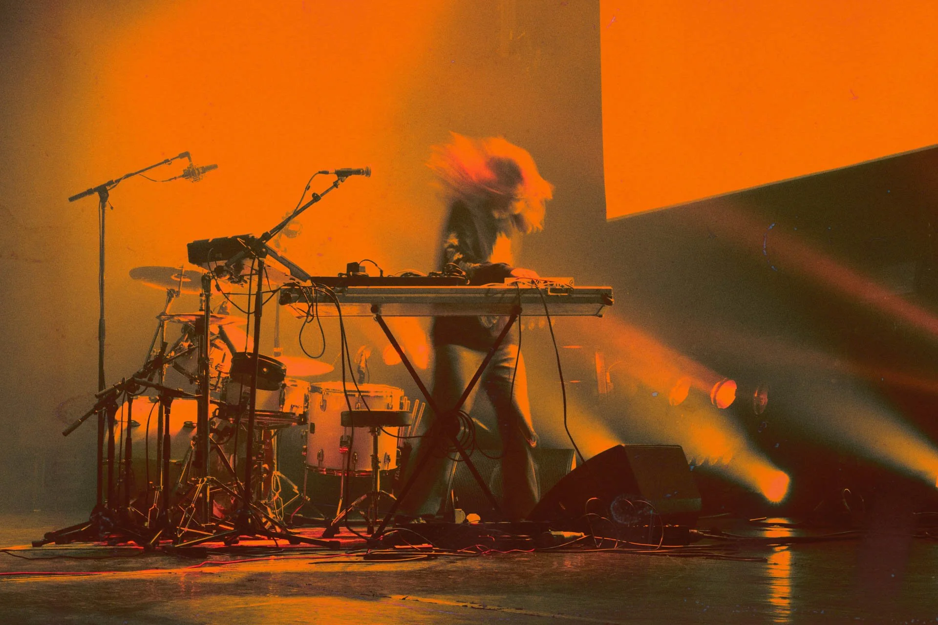 Musicien jouant du clavier sur scène, éclairage orange avec brume ou fumée, set de batterie en arrière-plan.