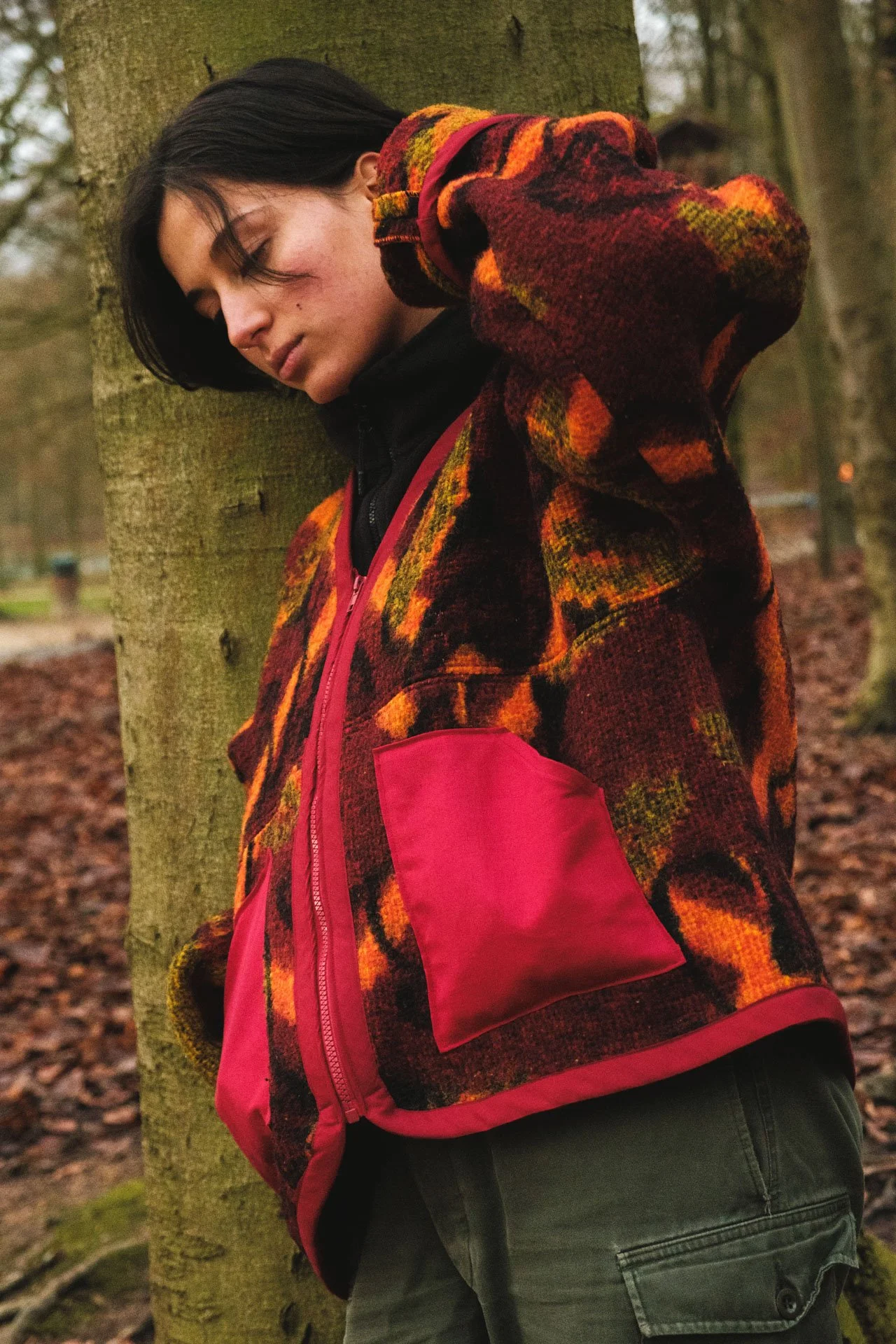 Jeune femme avec un manteau en laine à motifs colorés, portant un sac à dos rose, se tenant contre un arbre dans un parc en automne.