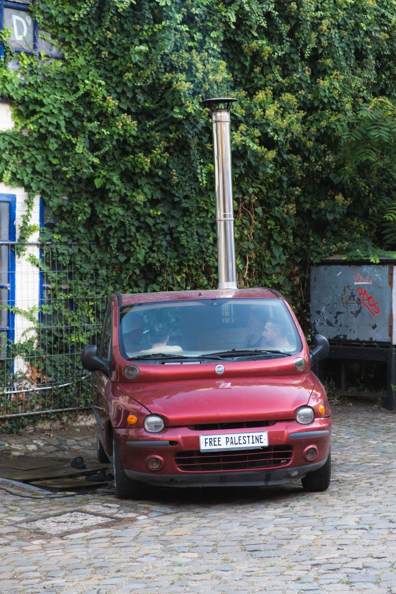 Une petite voiture rouge avec une plaque d'immatriculation indiquant 'FREE PALESTINE' est garée sur une surface pavée, devant un mur recouvert de végétation dense.