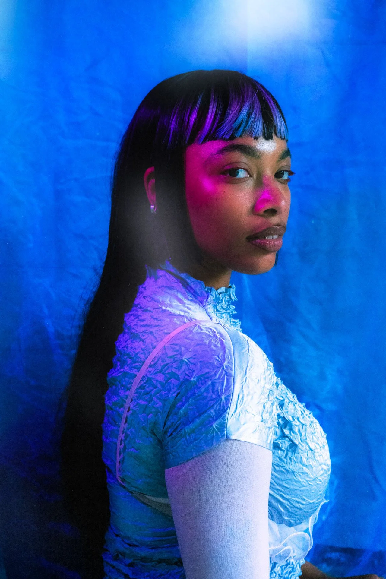Portrait d'une femme avec des cheveux noirs et décolorés en violet, bleuté et noir, portant une veste blanche texturée, sur un fond bleu violet lumineux.
