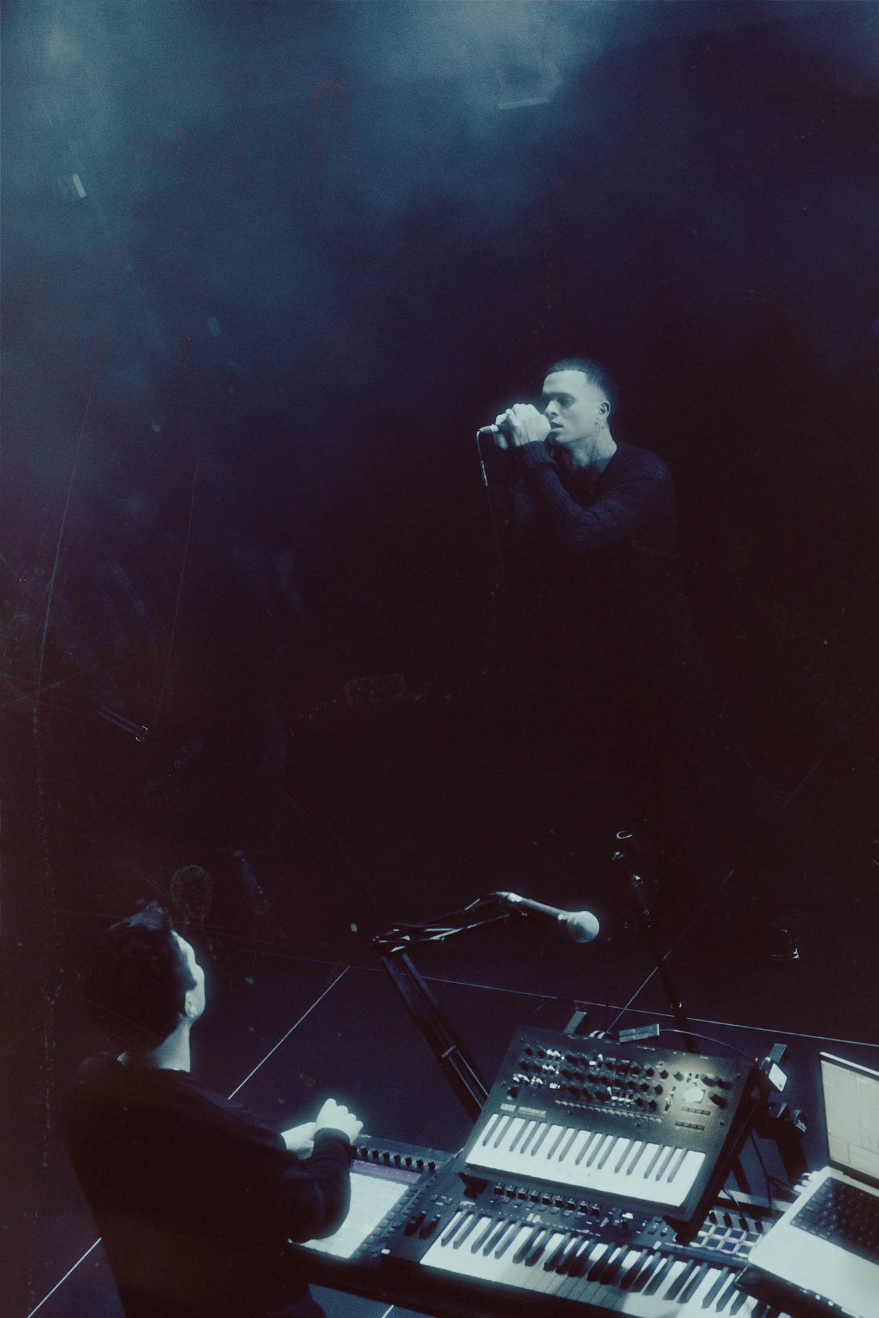 Un homme chante dans un microphone sur scène, avec un clavier, un synthétiseur et un contrôleur MIDI devant lui, dans une ambiance sombre avec une lumière bleue.