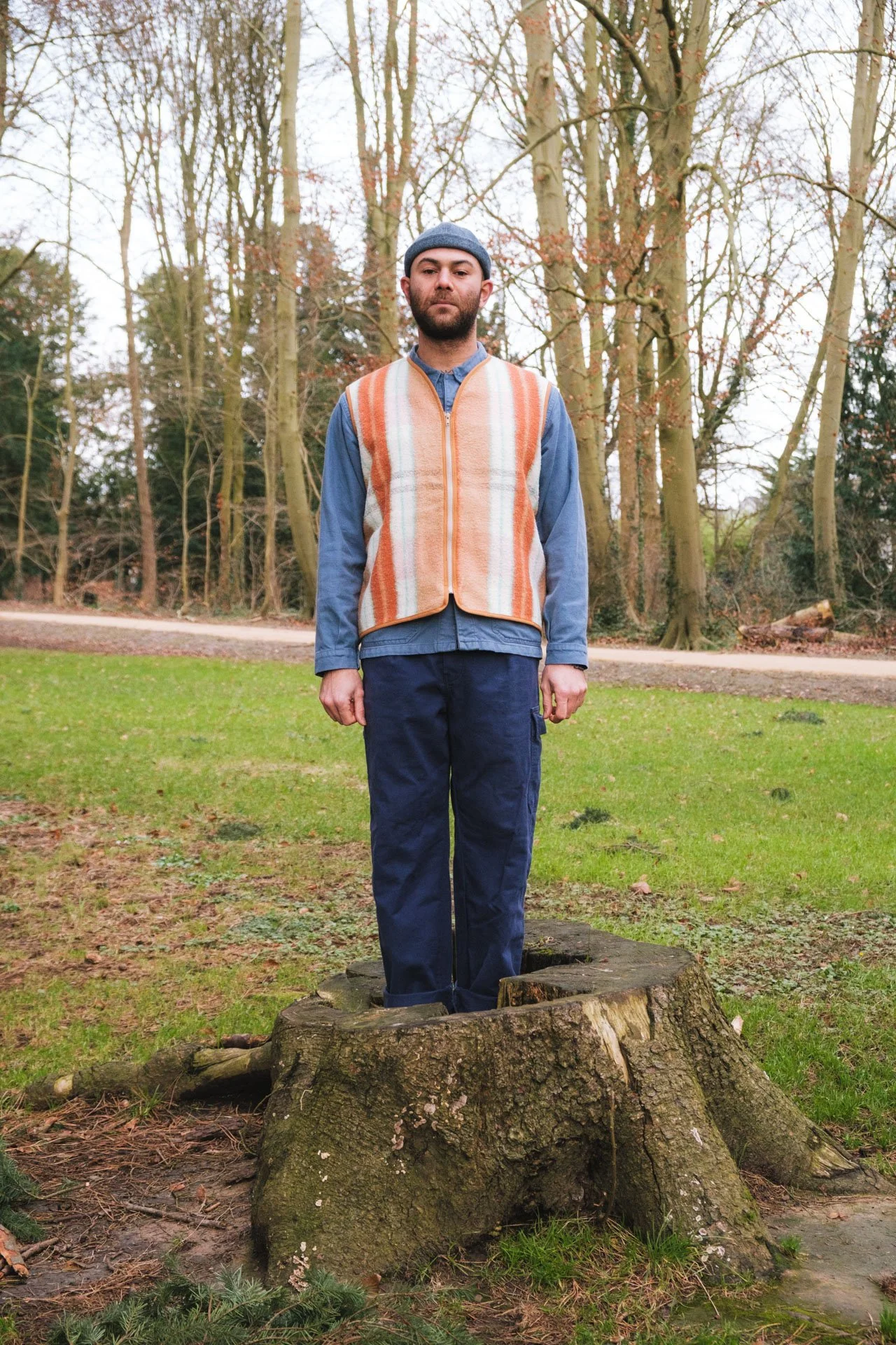 Un homme debout sur une souche d'arbre dans un parc, entouré d'arbres dénudés, vêtu d'une veste en maille rayée orange, d'une chemise bleue et d'un pantalon bleu, avec un bonnet gris