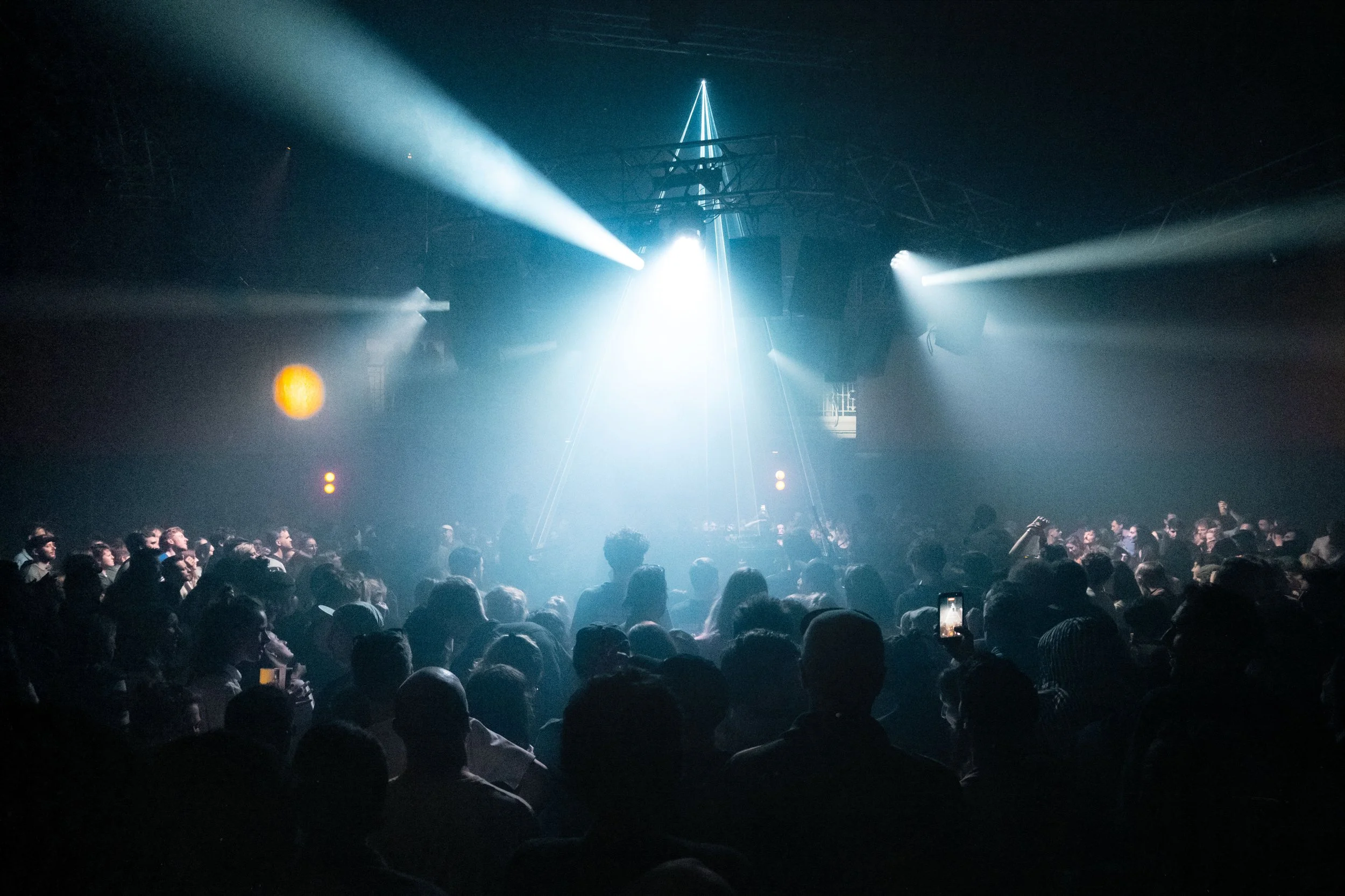 Une foule de personnes dans une salle de concert ou de spectacle sombre, avec des lumières colorées et des faisceaux lumineux dirigés vers le plafond.