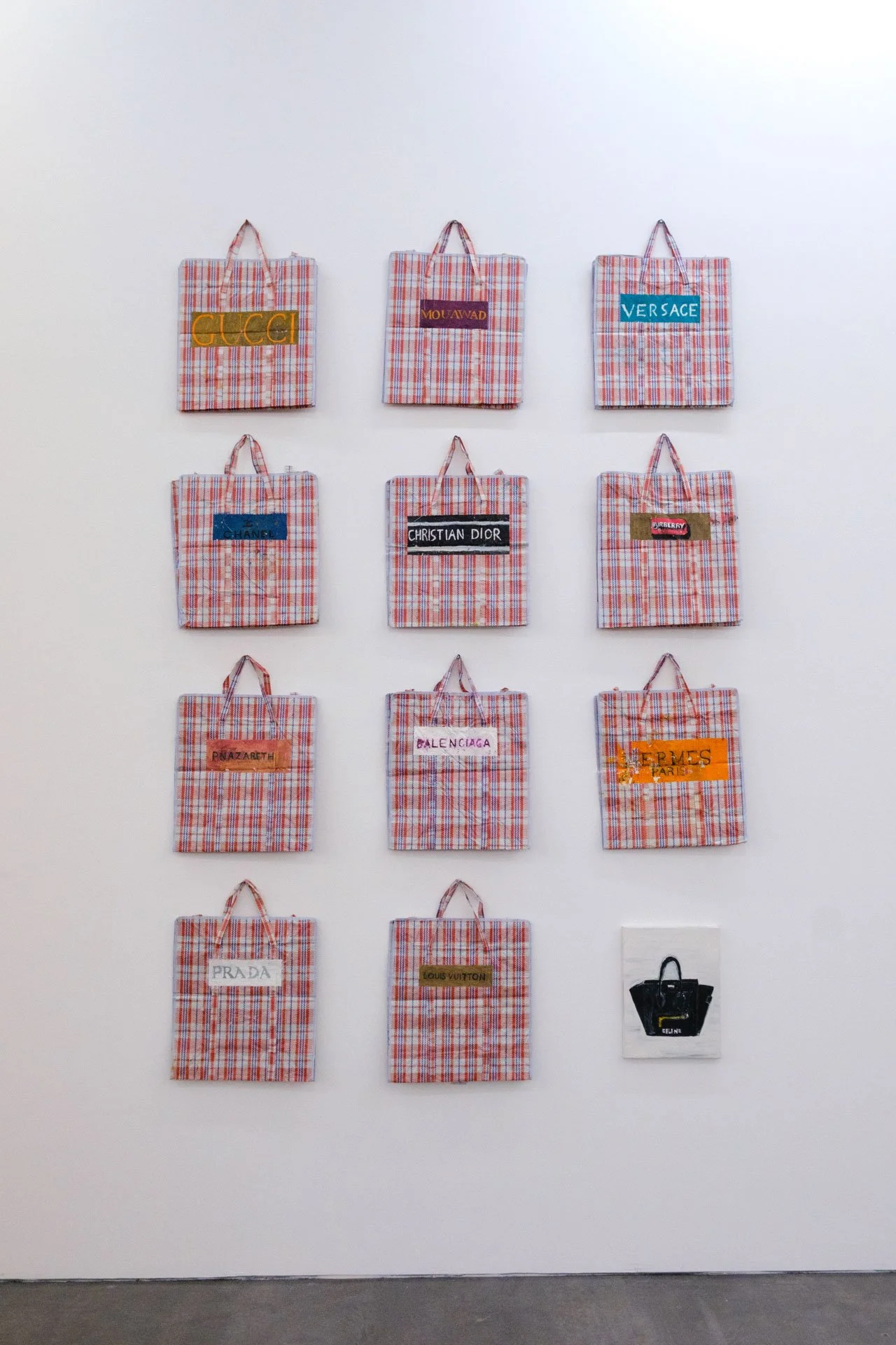 Une installation artistique avec des sacs à main de luxe célèbres de marques comme Gucci, Chanel, Christian Dior, Balenciaga, Prada, Louis Vuitton et Hermès, suspendus contre un mur blanc dans une galerie d'art.