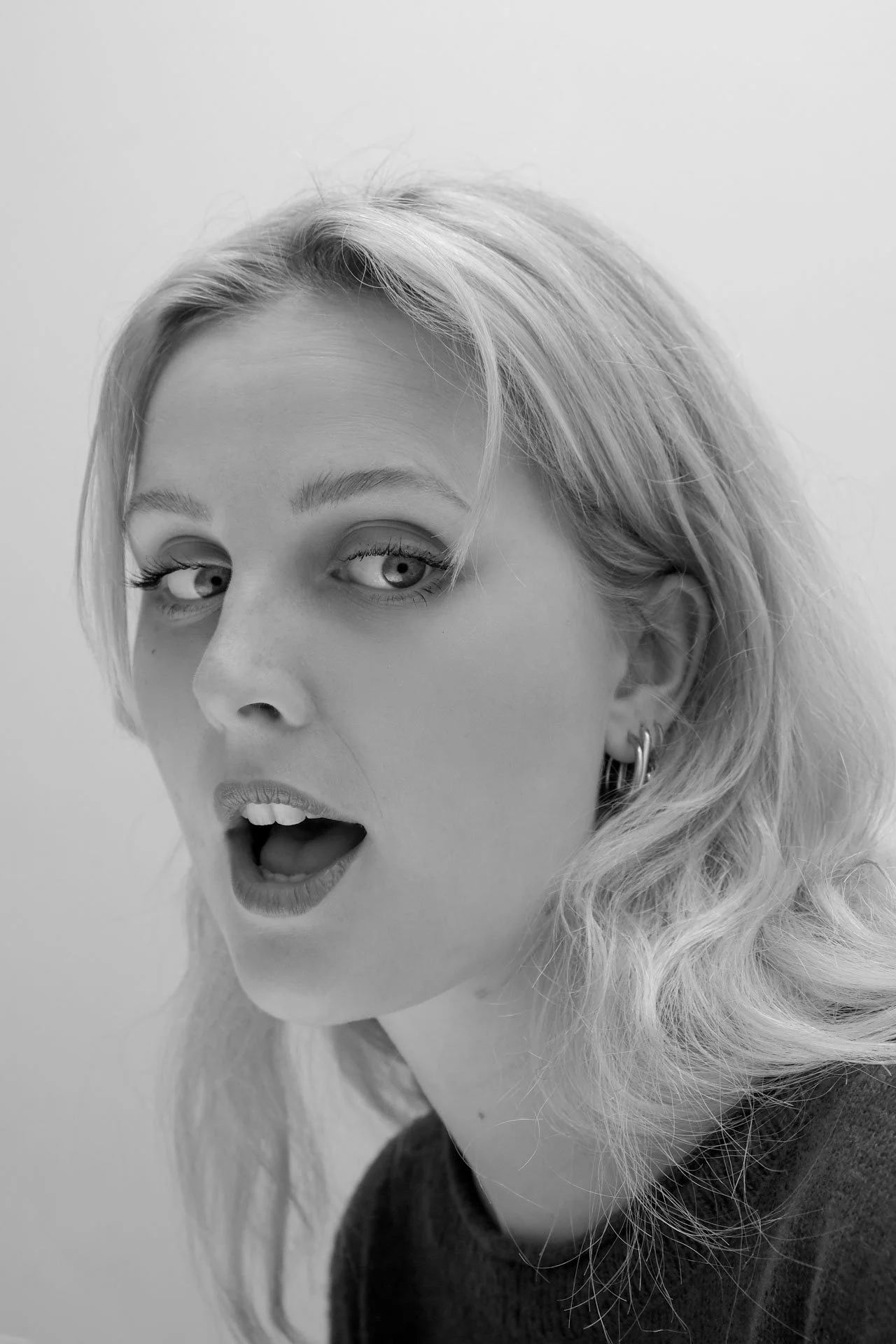 Jeune femme aux yeux bleus, yeux grands ouverts, exprimant une émotion de surprise ou d'émerveillement. Long cheveux blonds, portant des boucles d'oreilles, dans un décor simple en noir et blanc.