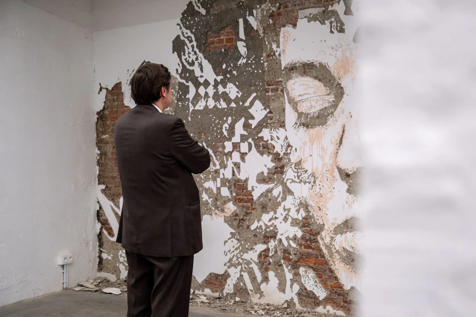 Un homme en costume regardant un mur en briques partiellement détruit, avec une oeuvre d'art représentant un portrait de femme, dans une galerie d'art.