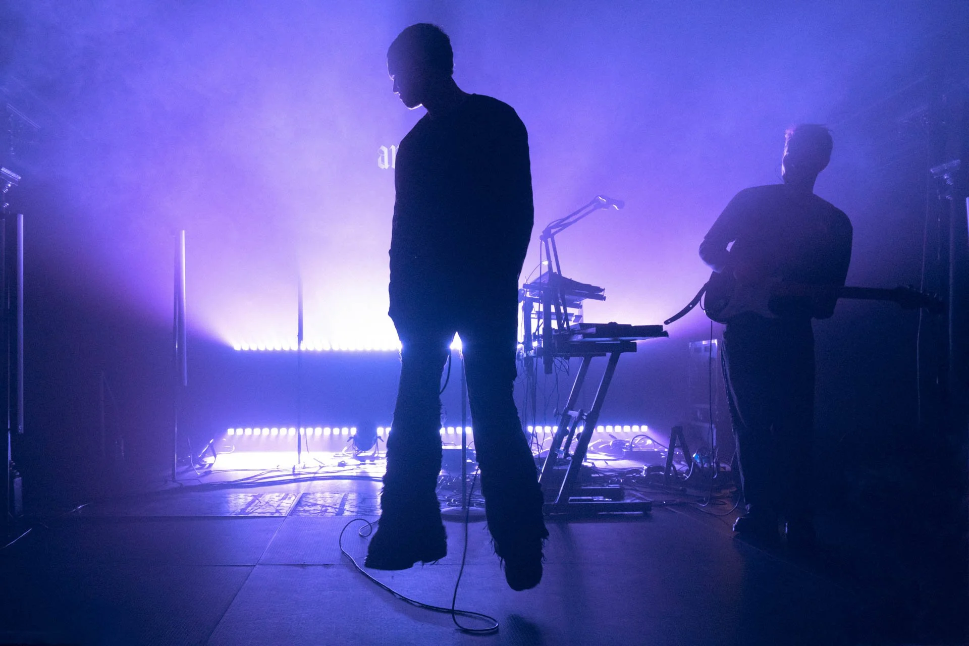 Deux musiciens silhouettes sur scène avec des lumières violettes et bleues, un à gauche avec un instrument de musique et un à droite avec une guitare, équipements de musique en arrière-plan.