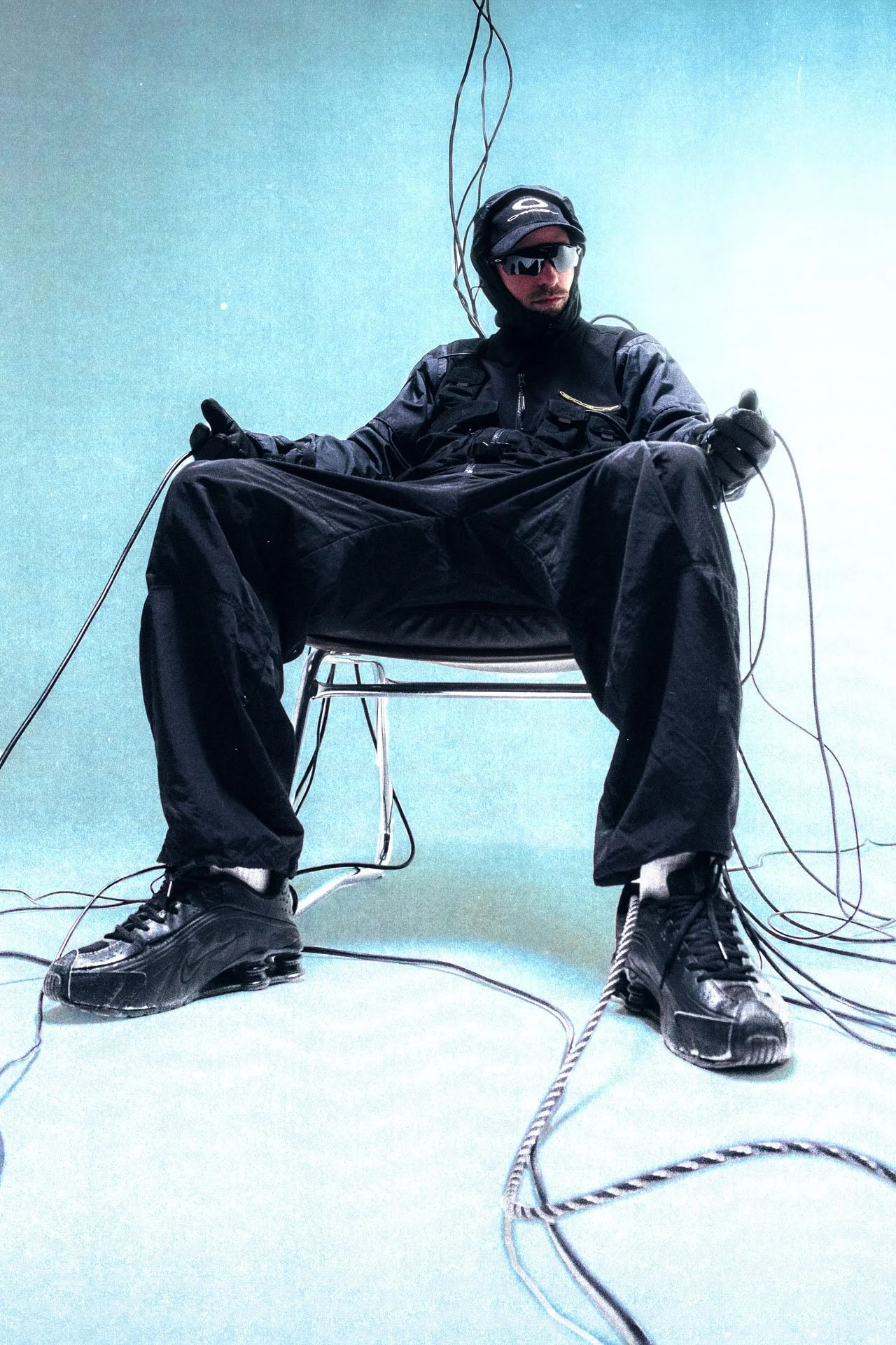 Une personne portant des vêtements noirs, des lunettes de soleil et un casque, assise sur une chaise avec des câbles électriques autour d'elle, dans un environnement bleu clair.