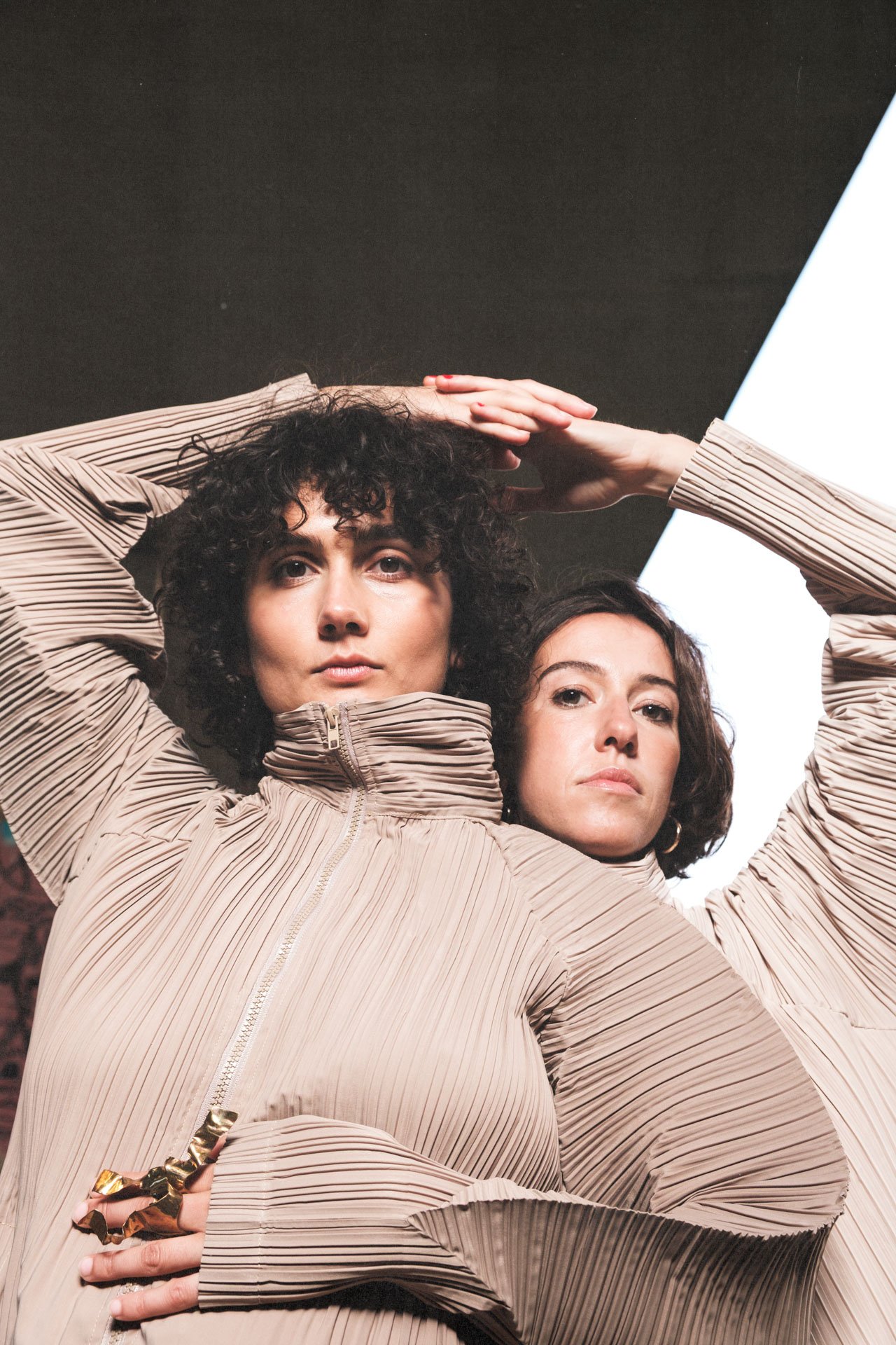 Deux femmes posent sous un éclairage moderne, portant des vestes beige avec des plis urilés pour une séance photo mode.