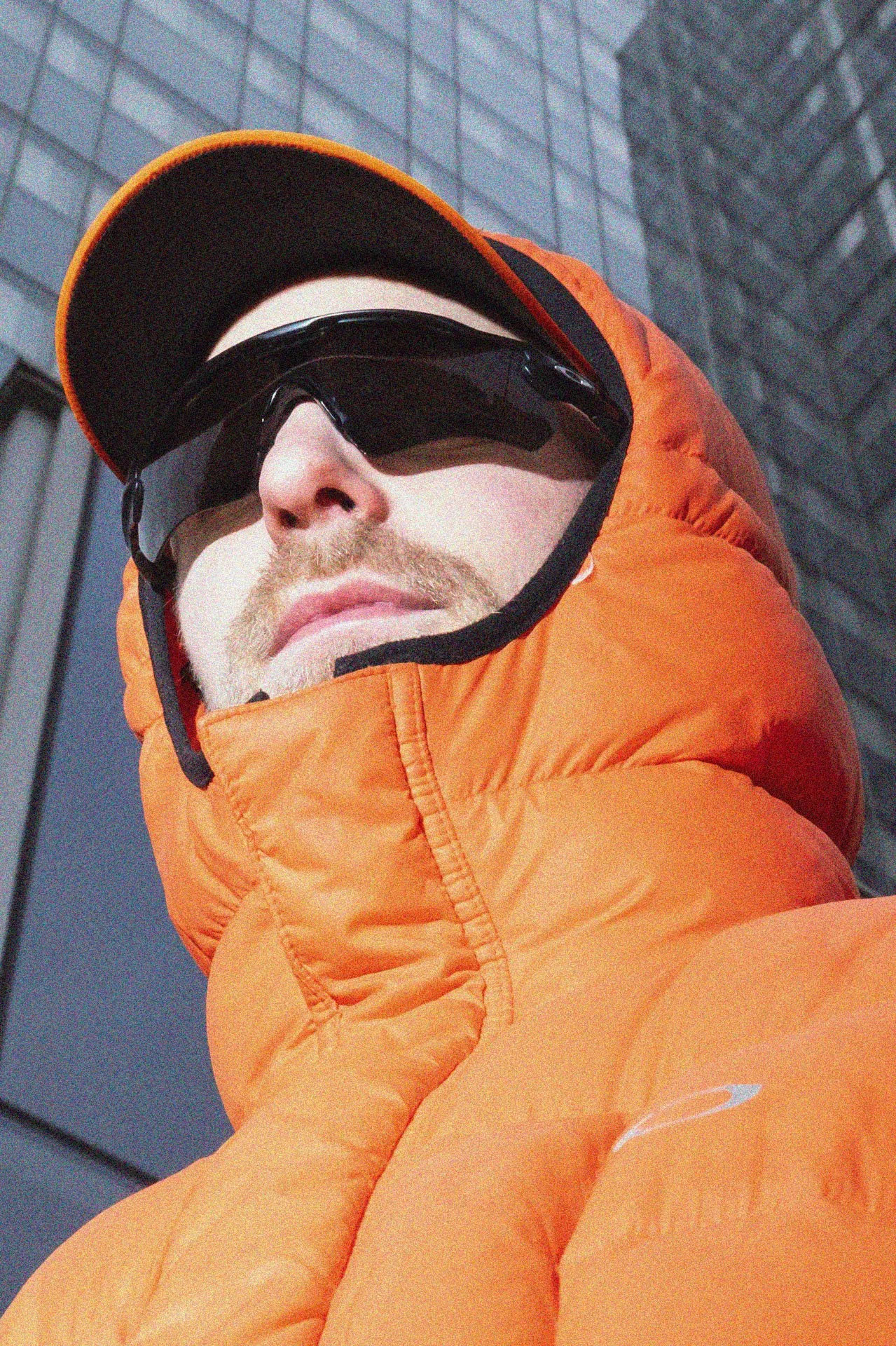 Un homme portant une casquette, des lunettes de soleil noires, un manteau orange avec capuche et un fond de mur en briques grises.