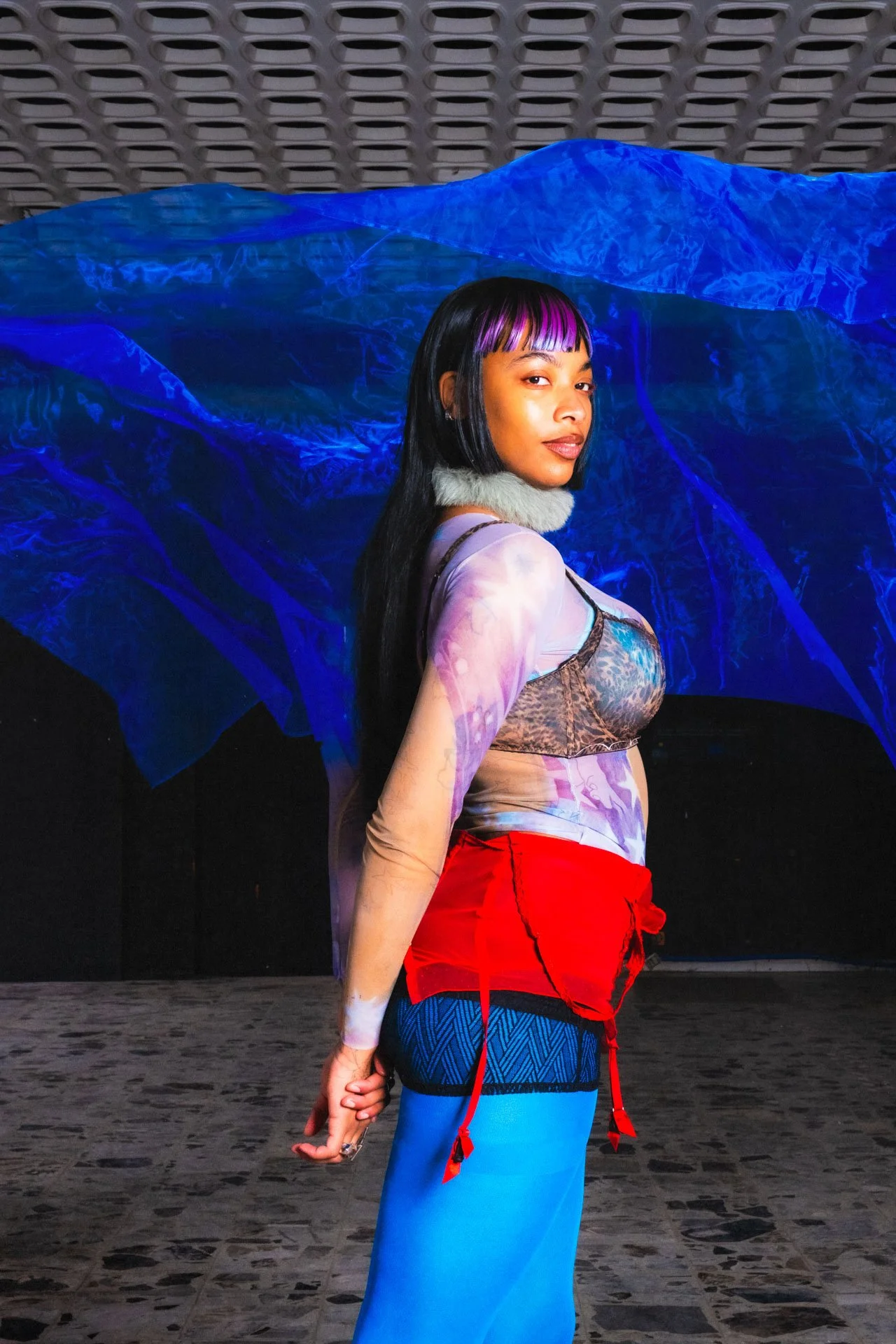 Une femme posant devant une toile d'art avec un fond lumineux bleu, portant une tenue colorée avec des manches translucides et une jupe rouge.