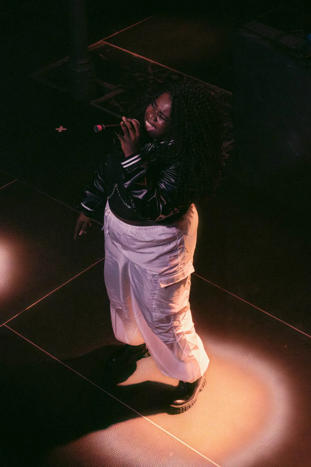 Une femme chante dans une ambiance sombre, portant un blouson en cuir noir et un pantalon ample blanc, avec des vêtements décontractés et une coiffure bouclée. Elle tient un microphone et semble exprimer une émotion intense.
