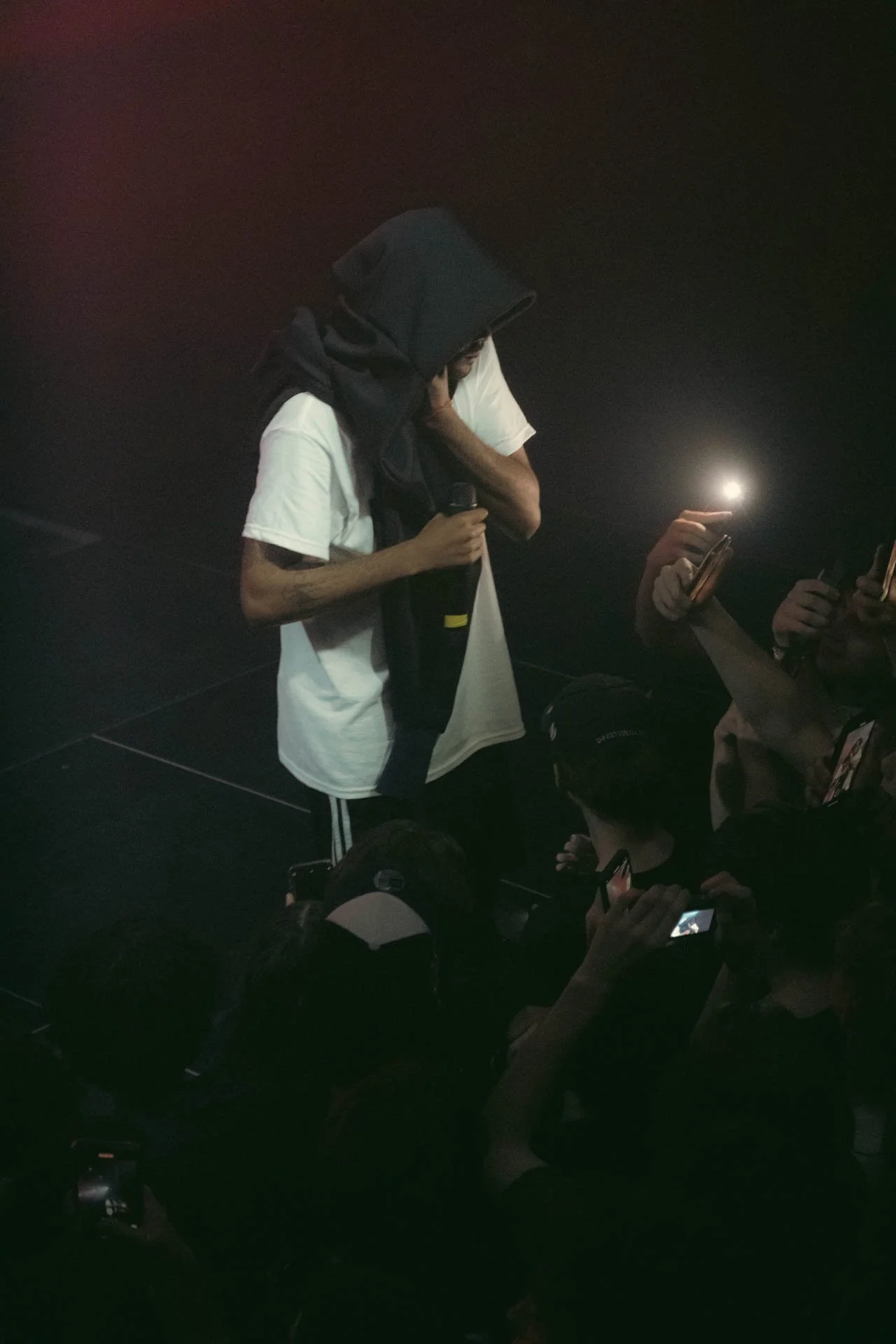 Jeune homme avec un hoodie noir et un t-shirt blanc, tenant un microphone, faisant signe avec la main à des personnes qui prennent des photos ou vidéos, dans un environnement sombre.