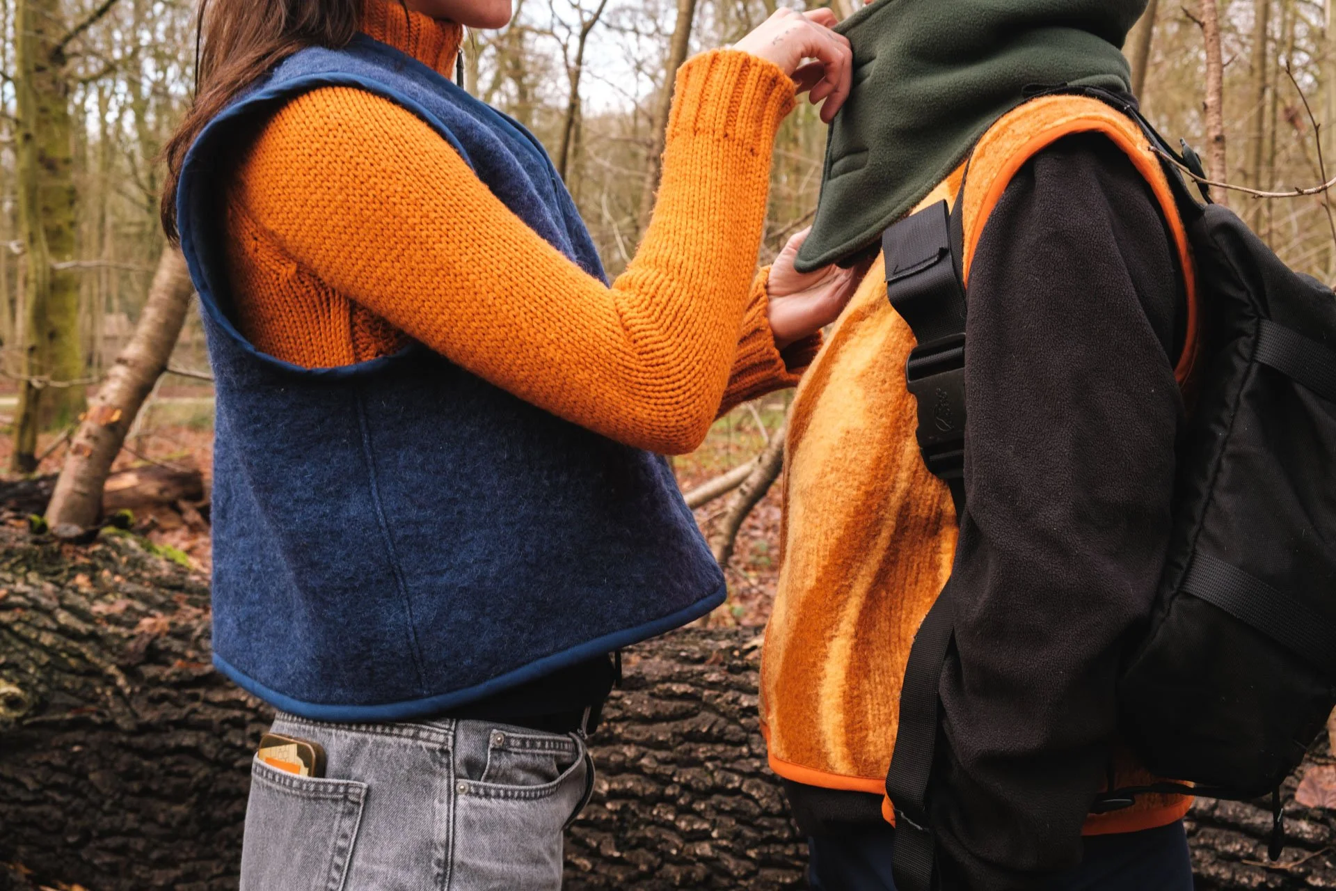 Deux personnes dans une forêt, l'une en veste orange et la petite en veste verte, en train d'interagir, probablement une conversation ou un contact tactile.