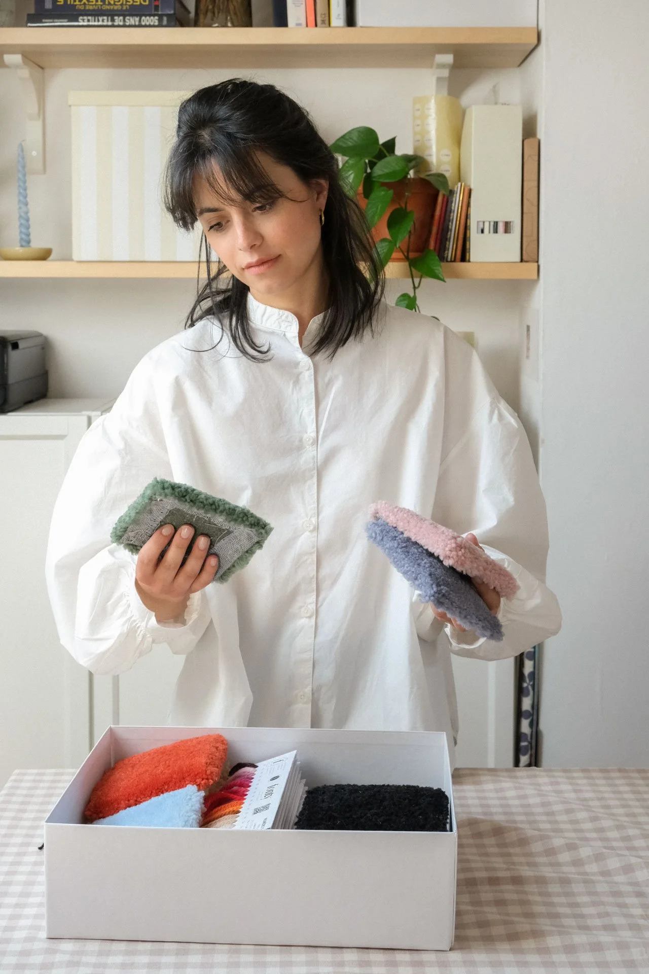 Une femme regarde des échantillons de textiles de couleur différente dans une boîte en carton sur une table, dans une pièce avec étagères contenant des livres, une plante et des objets décoratifs.
