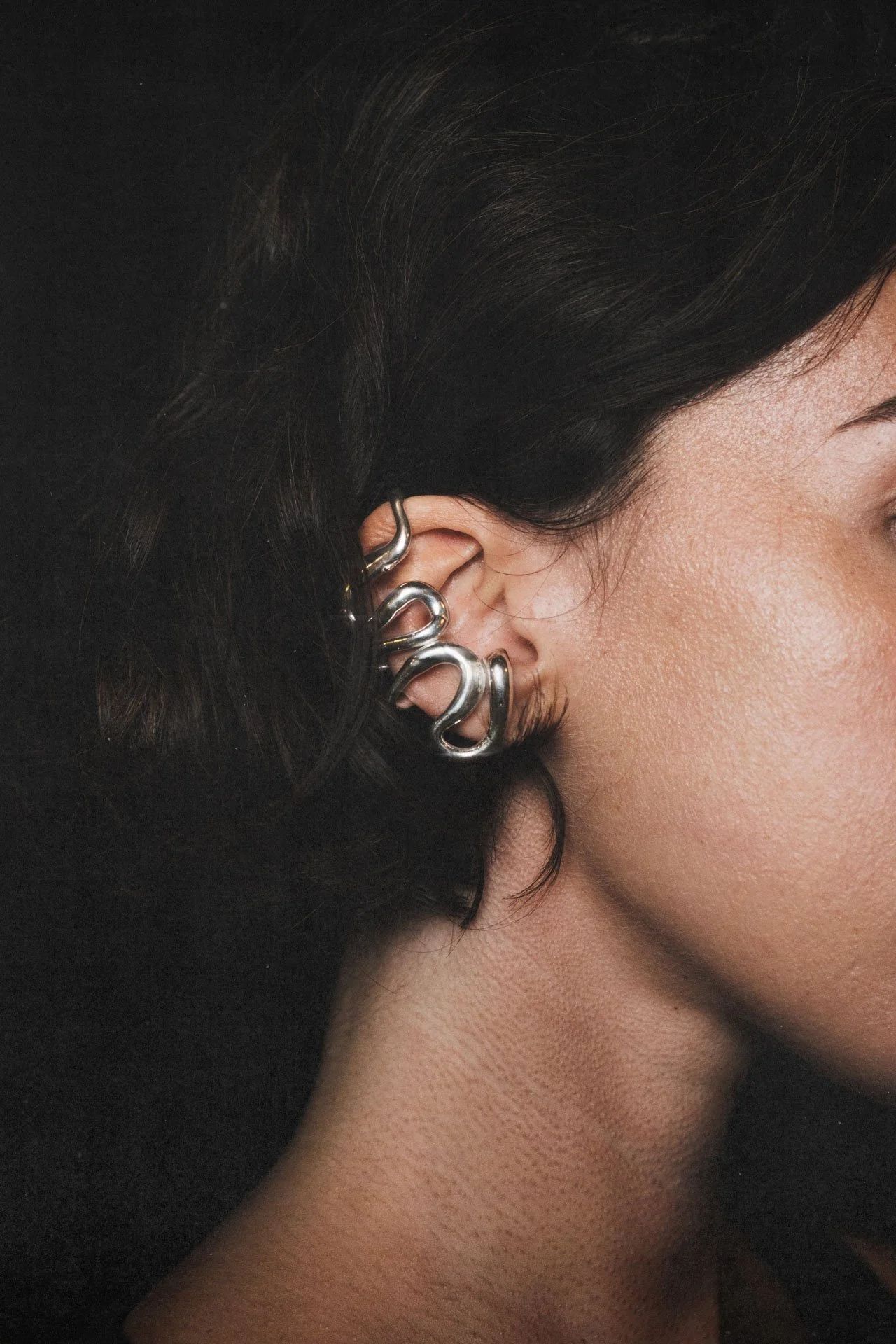 Un gros plan d'une oreille ornée d'une boucle d'oreille en métal argenté avec un design ondulé, avec des cheveux sombres et un visage partiellement visible à droite.
