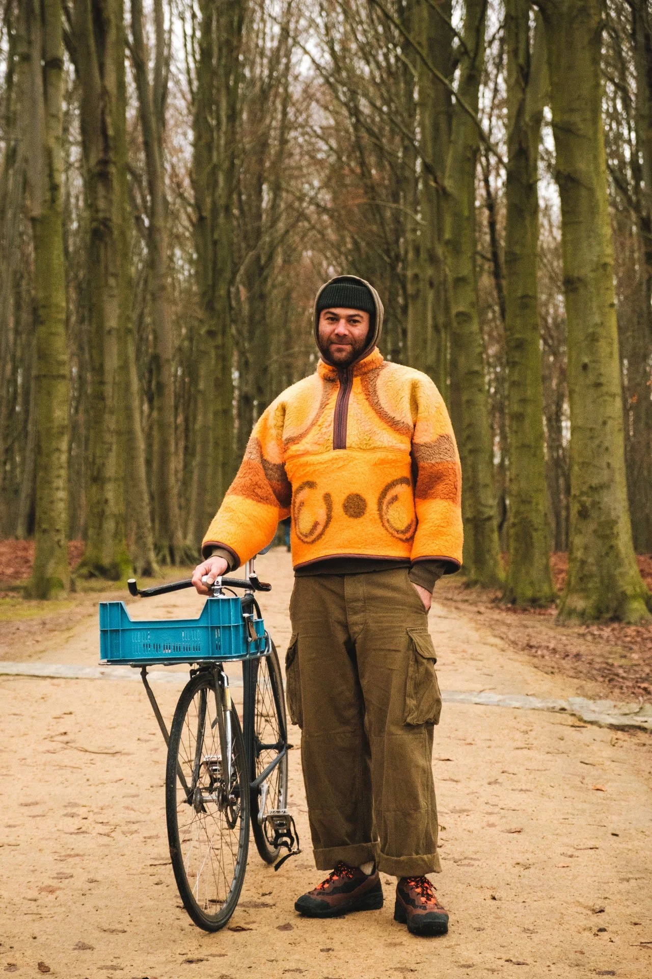 Un homme debout dans un parc, tenant un vélo avec un panier bleu, portant un pull orange avec un motif de visage souriant, des pantalons cargo kaki, et un bonnet noir, entouré d'arbres sans feuilles.