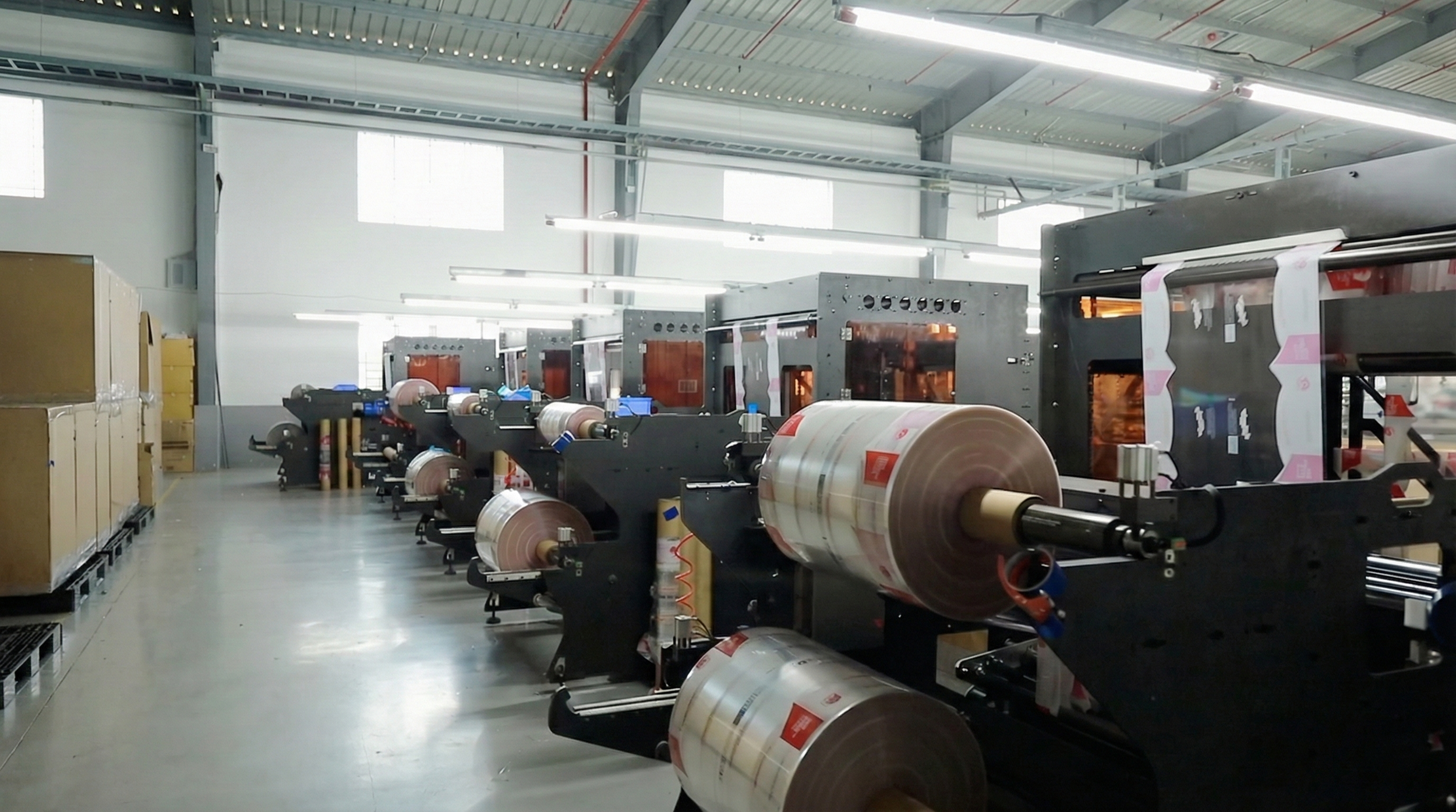 Industrial printing facility with large machines processing rolls of printed material, metal and concrete floors, high ceiling, fluorescent lighting.