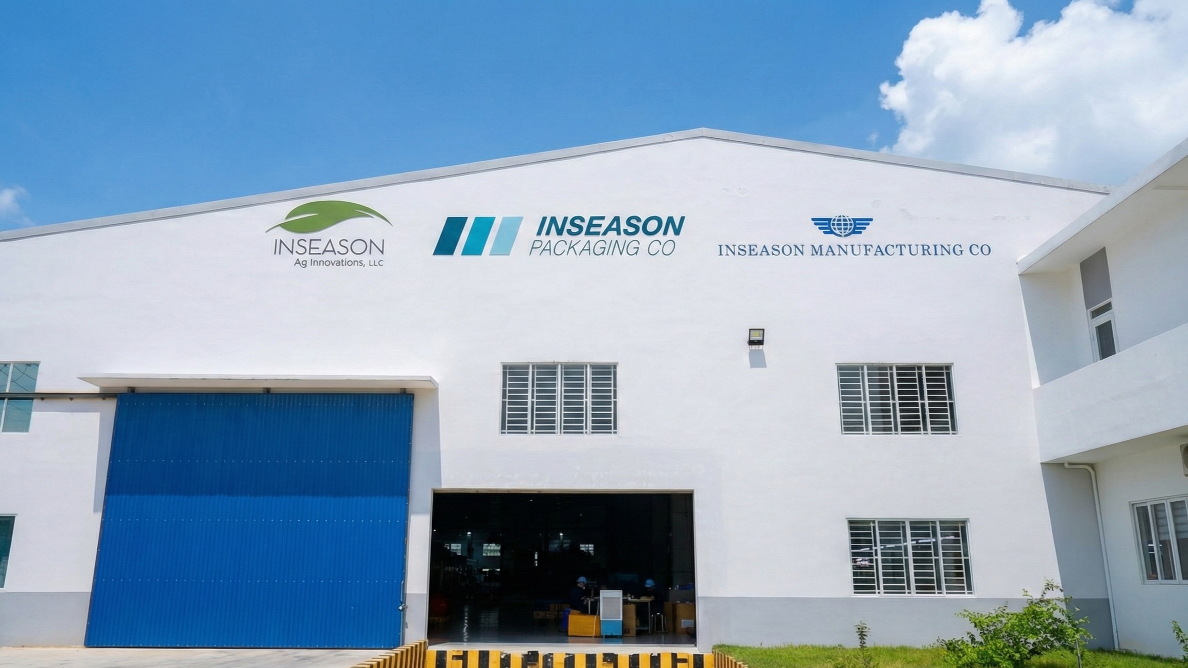 Front view of a white industrial building with logos of INSEASON Ag Innovations, LLC, INSEASON Packaging Co, and INSEASON Manufacturing Co on the facade, under a partly cloudy blue sky.