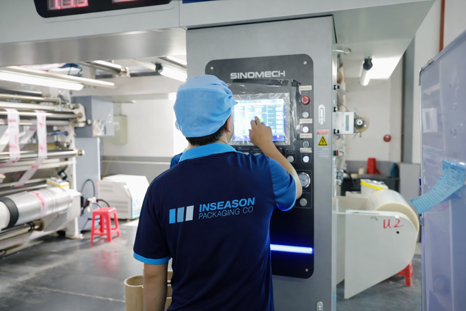 A worker from INSEASON PACKAGING CO wearing a blue uniform and cap operating a printing or packaging machine in a factory.