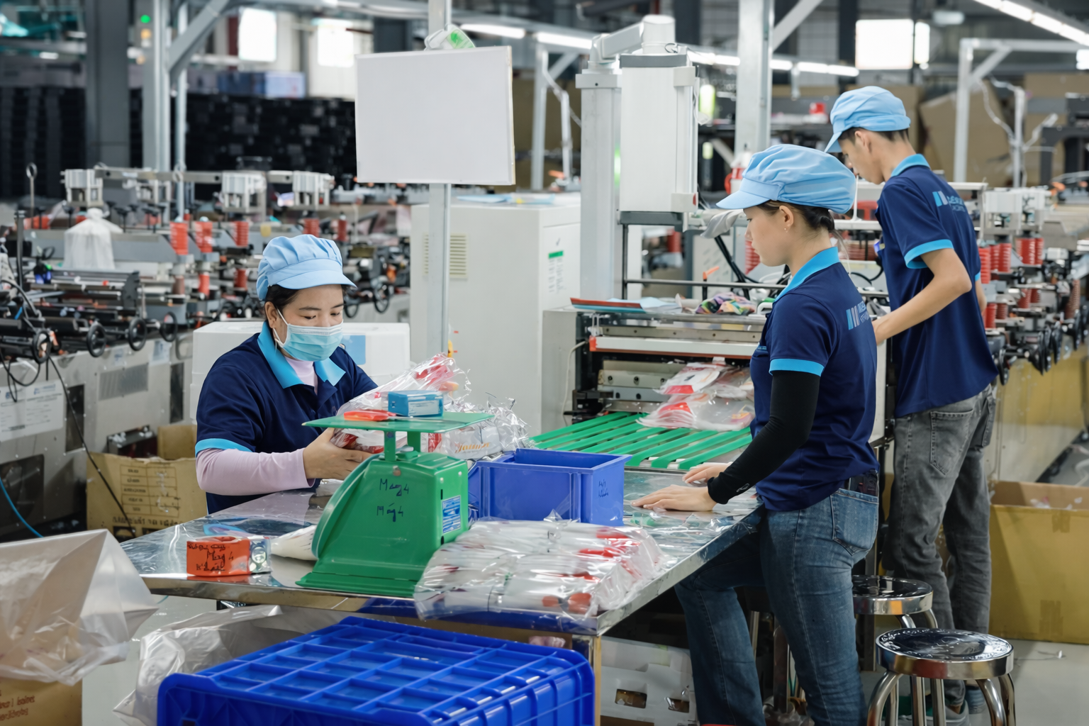 Workers in a factory assembling or packaging products at a work table, with machinery and boxes in the background.