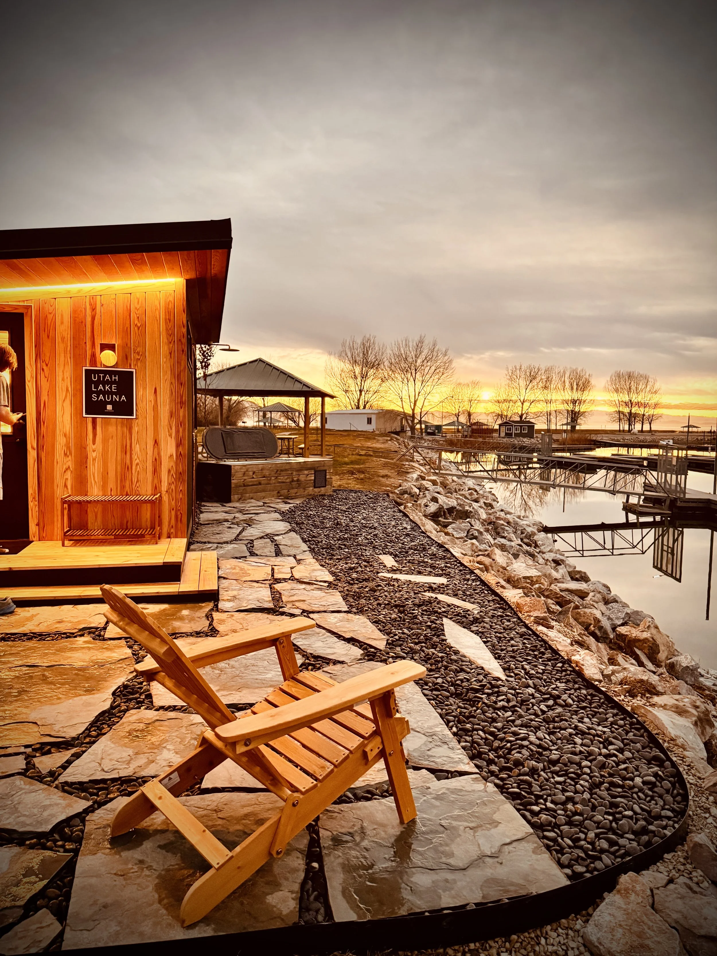 Sunset view with a wooden Adirondack chair near a small sauna labeled 'Utah Lake Sauna' by a lakeside, with trees, docks, and calm water.