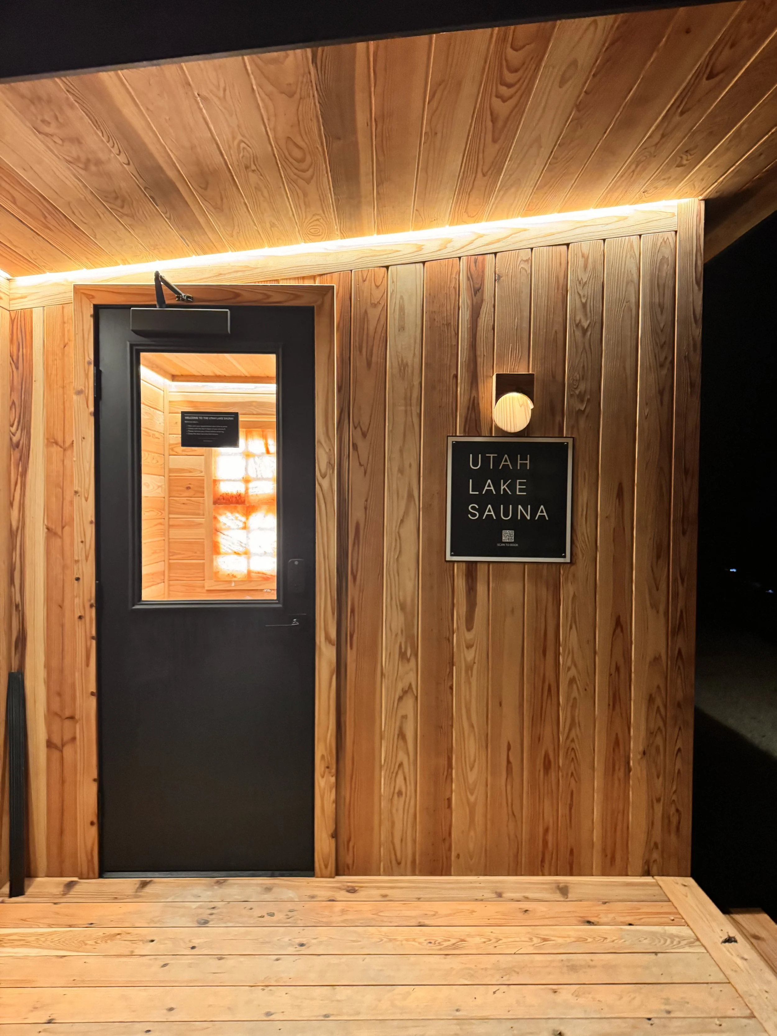 Wood-paneled entrance to Utah Lake Sauna with a black door, sign, and warm lighting.