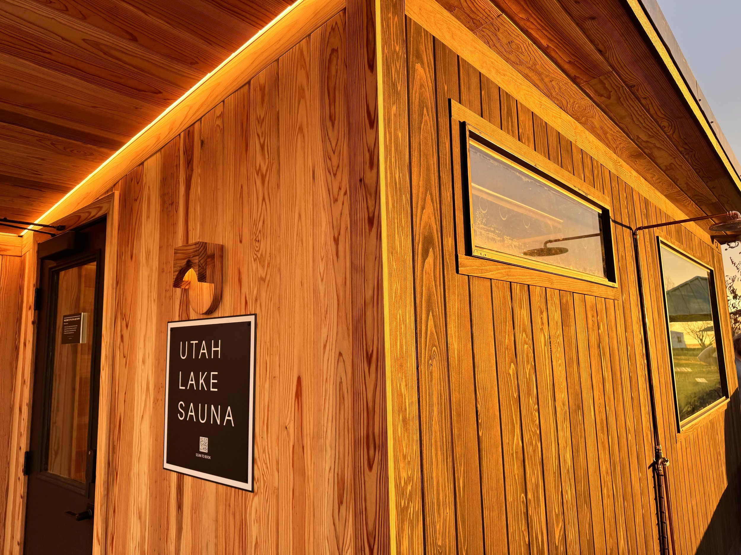 Exterior of a wooden building with a sign that reads 'Utah Lake Sauna' and small horizontal windows, illuminated by warm yellow lighting.