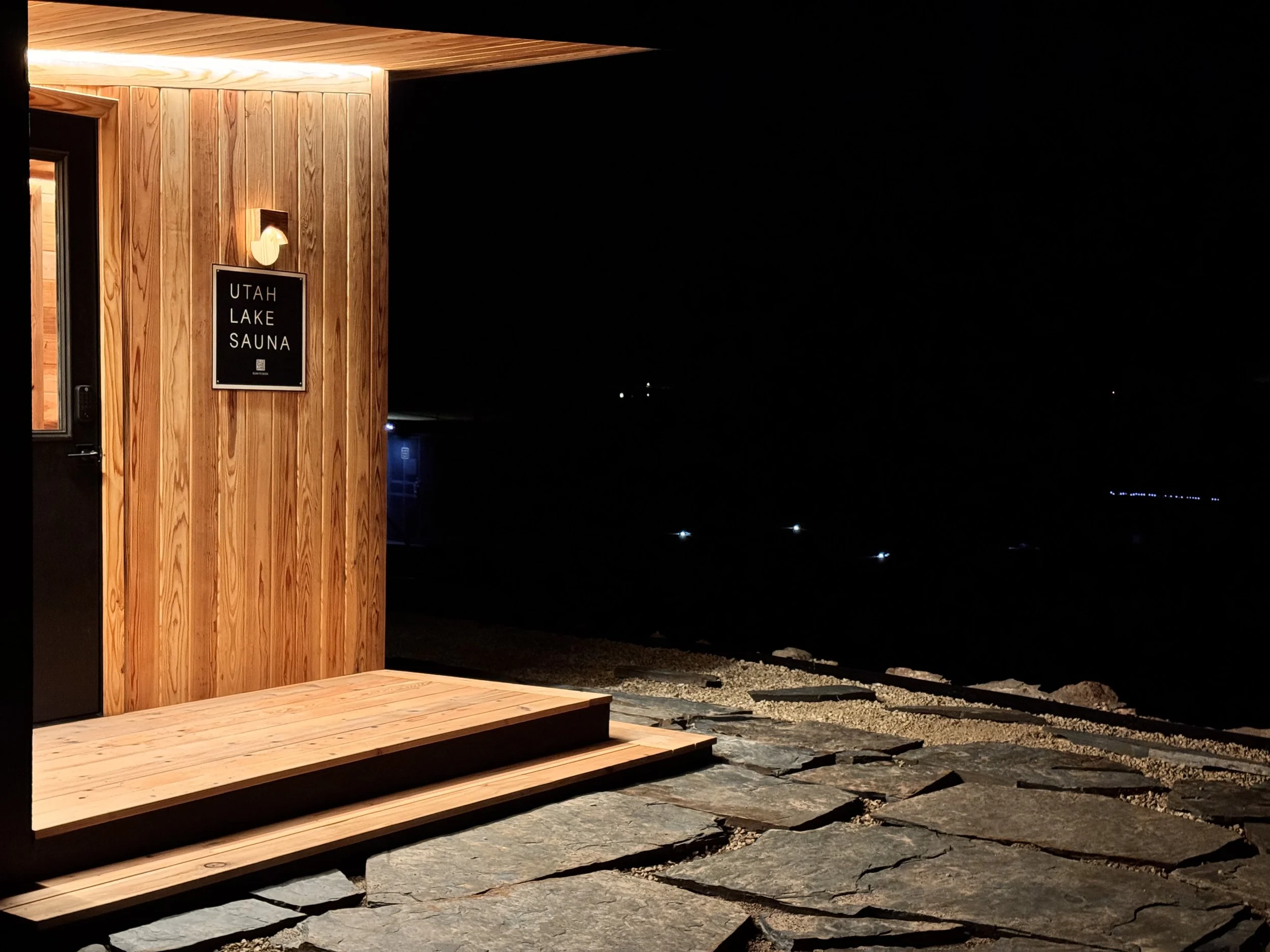 Nighttime view of the entrance to Utah Lake Sauna, with a wooden exterior and steps leading up to a door, overlooking a dark lake with distant lights.